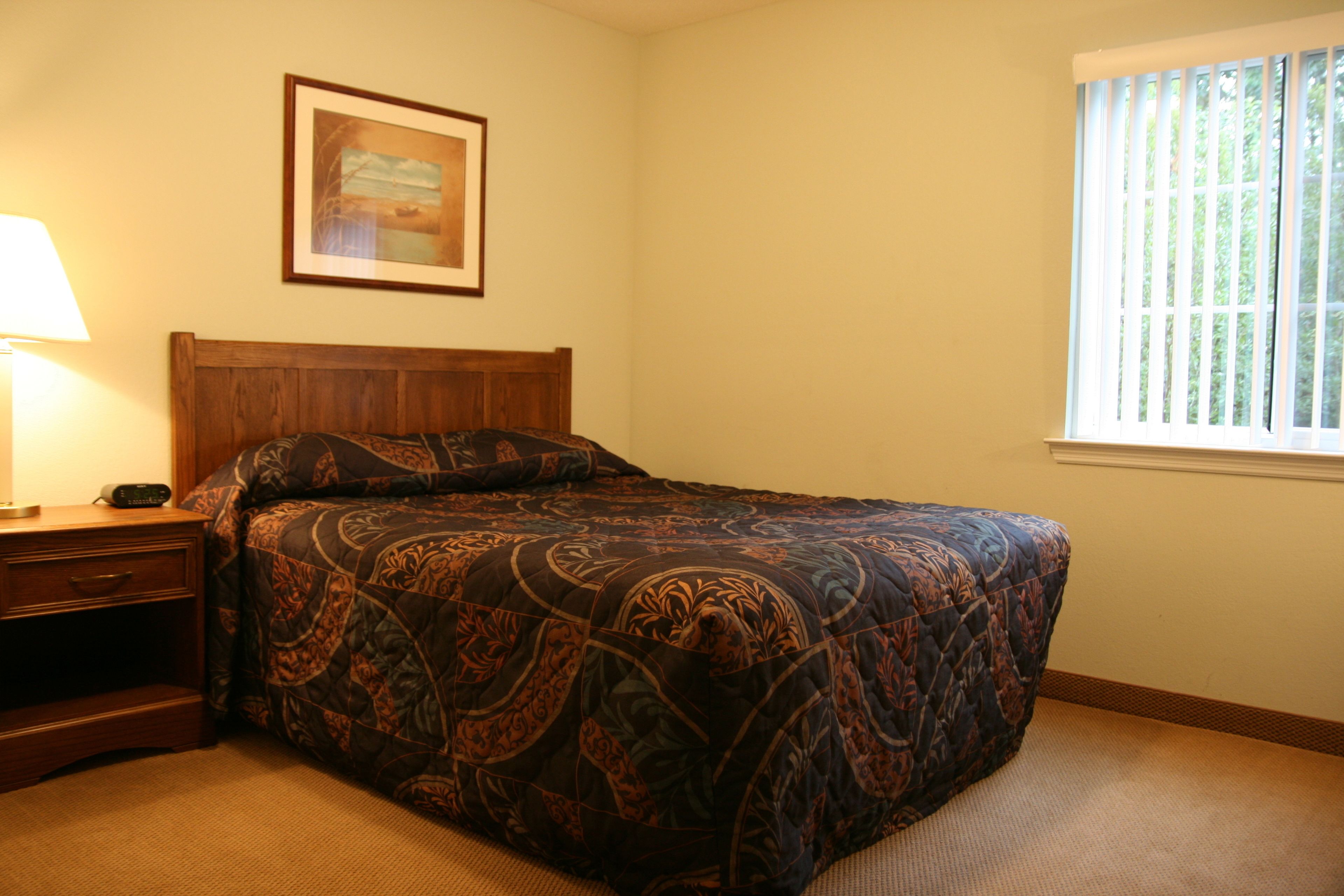 Une chambre à coucher avec un lit, une table de chevet, une lampe, un tableau encadré au mur et une fenêtre avec des stores.