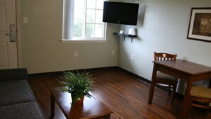Ein Wohnzimmer mit Holzboden, einer Couch, einem Tisch mit einer Pflanze, einem an der Wand befestigten Fernseher und einem Sessel.