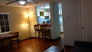 Ein Zimmer mit Holzboden, einem Esstisch mit Stühlen, einem Deckenventilator und einer durchgehenden Tür erkennbaren Küchenzeile.