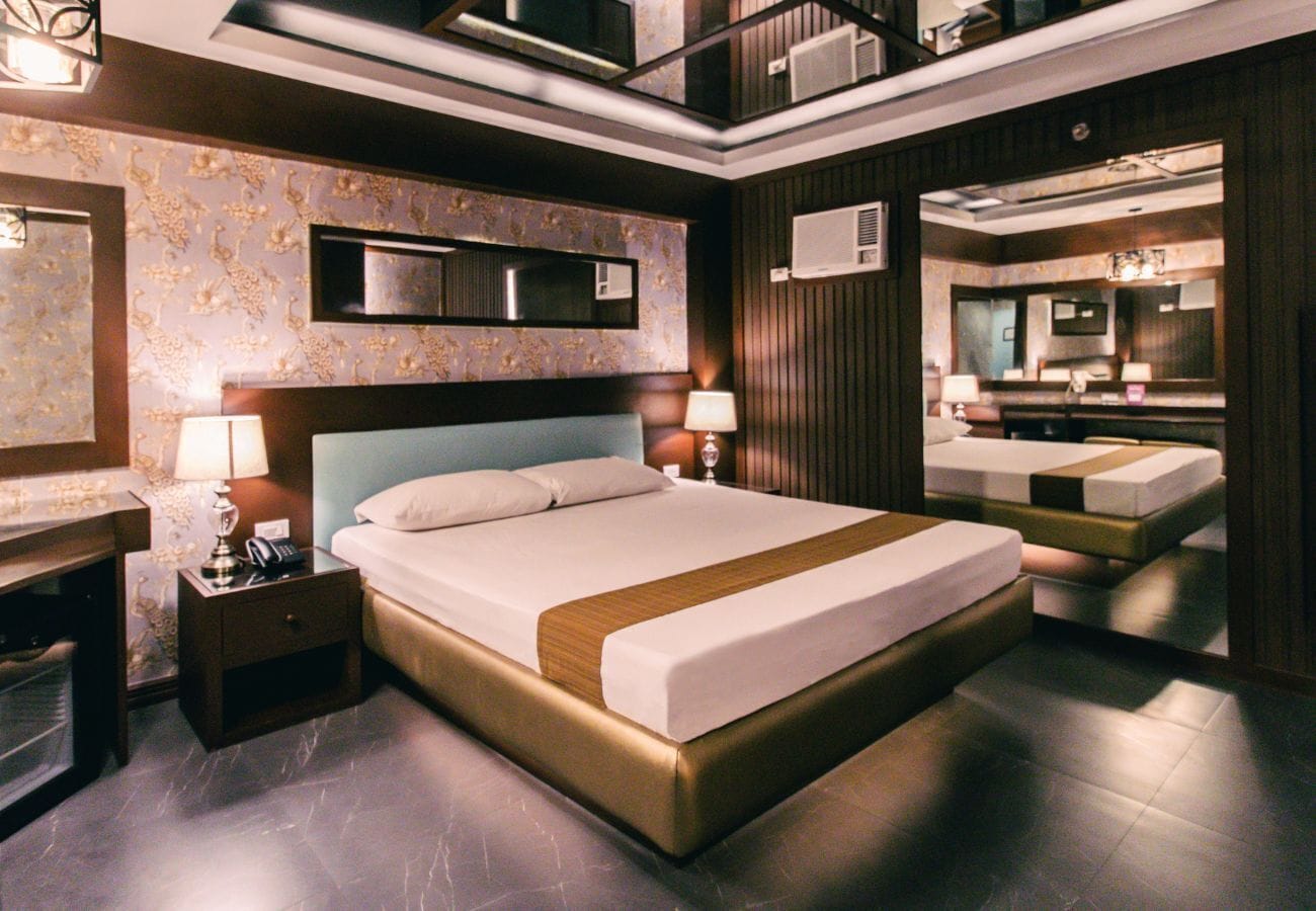 A modern hotel room with a large bed, a bedside table, a lamp, a mirror, and a wardrobe.