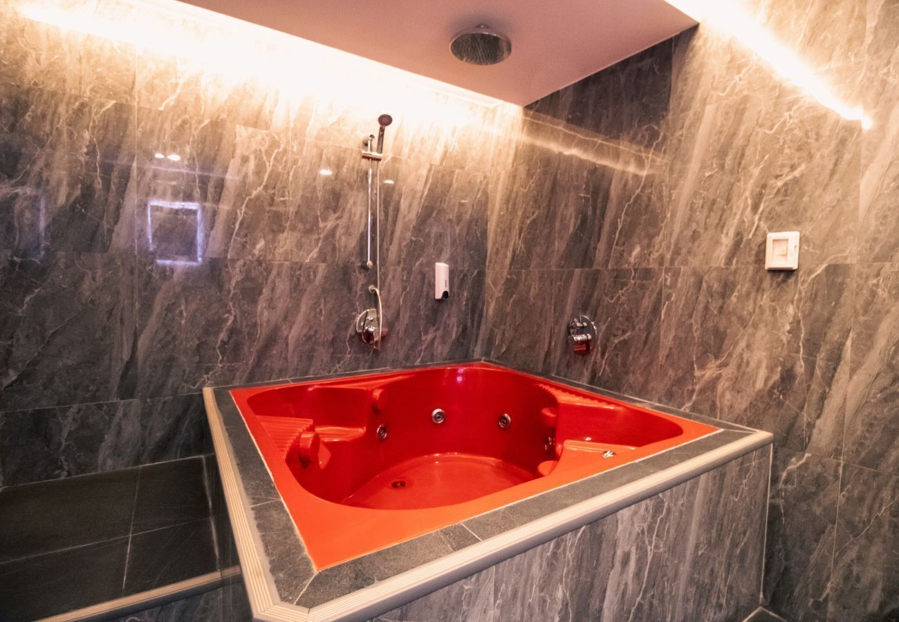 A modern bathroom with a large rectangular jacuzzi tub against a marble wall, a showerhead, and a small square mirror.