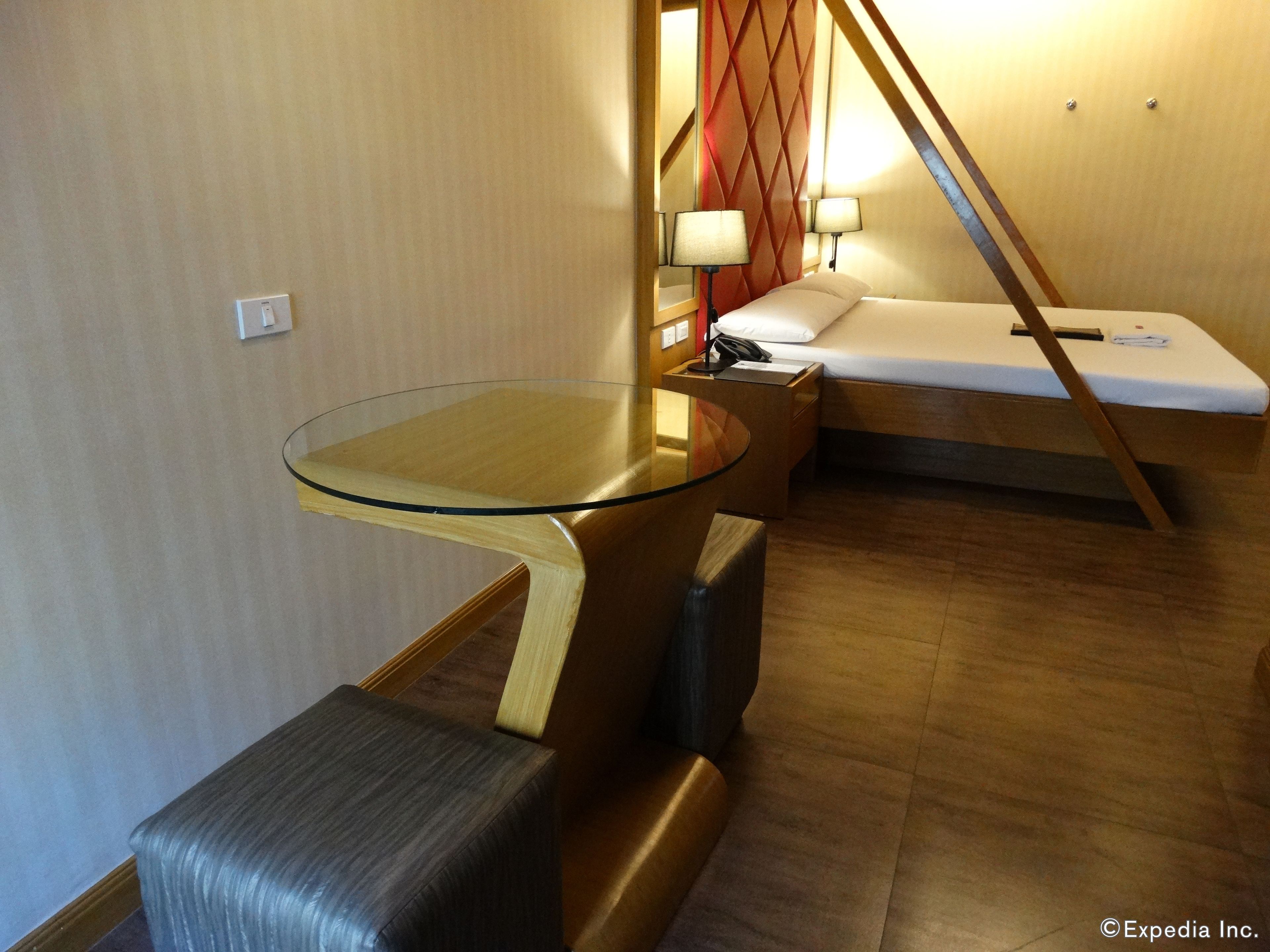 Ein kompaktes Hotelzimmer mit Glastisch, gepolstertem Sitzbänklein, einem Einzelbett und einem kleinen Nachttisch mit Lampen.