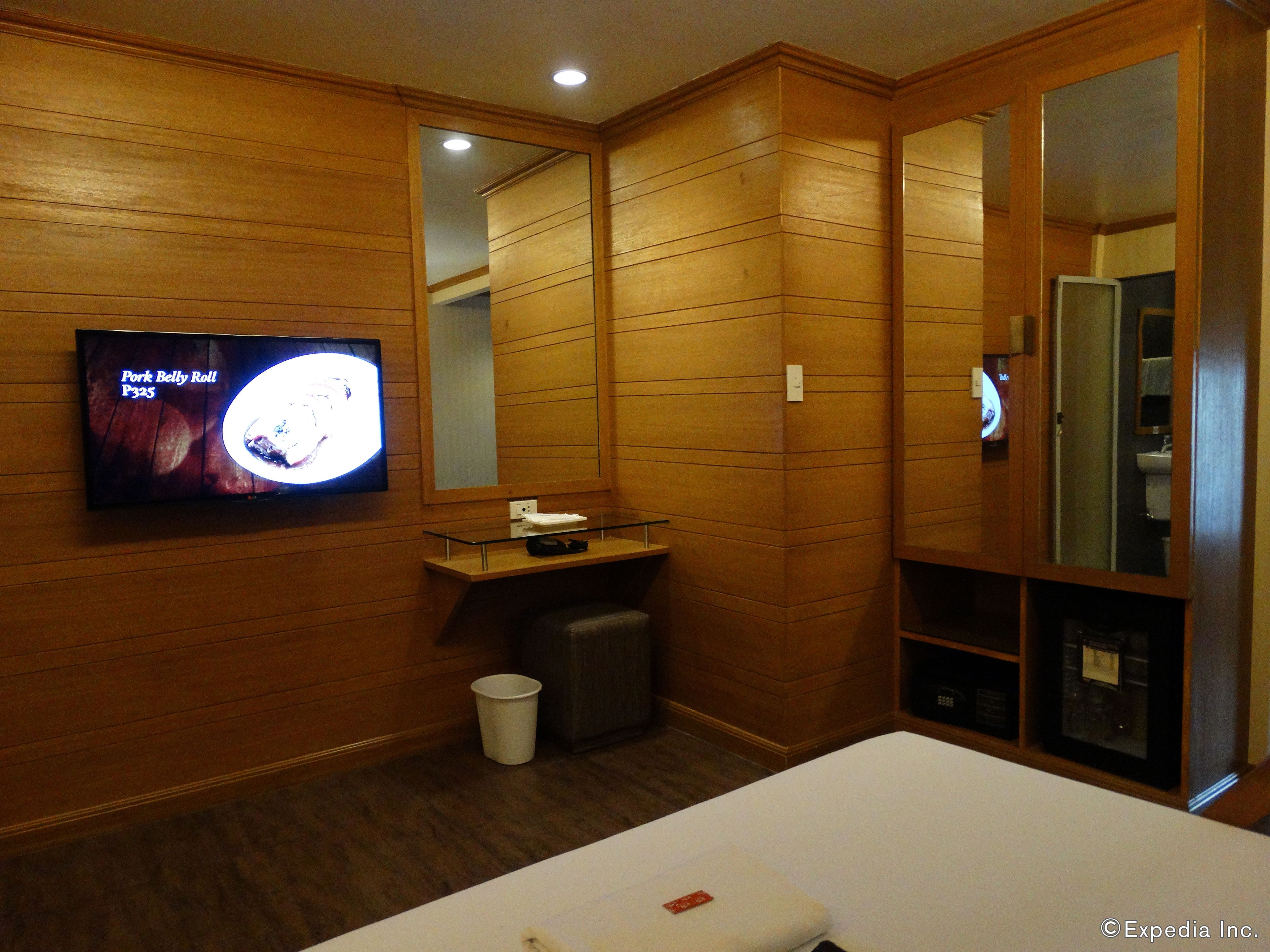 A hotel room with wooden paneling, a television displaying a menu, a small table with a glass top, a trash bin, and a bed with a white headboard.