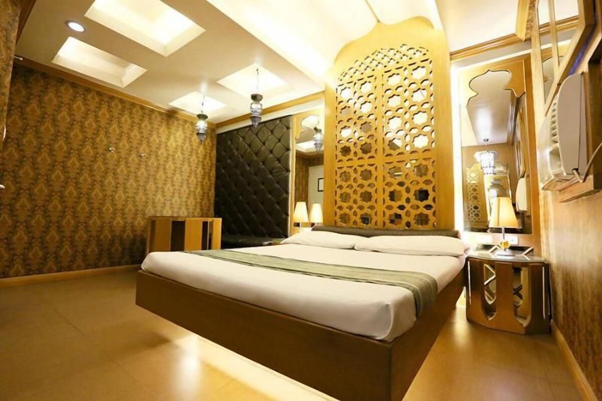 Habitación de hotel con una cama grande, un panel decorativo en la pared, un espejo y una mesita de noche con una lámpara.