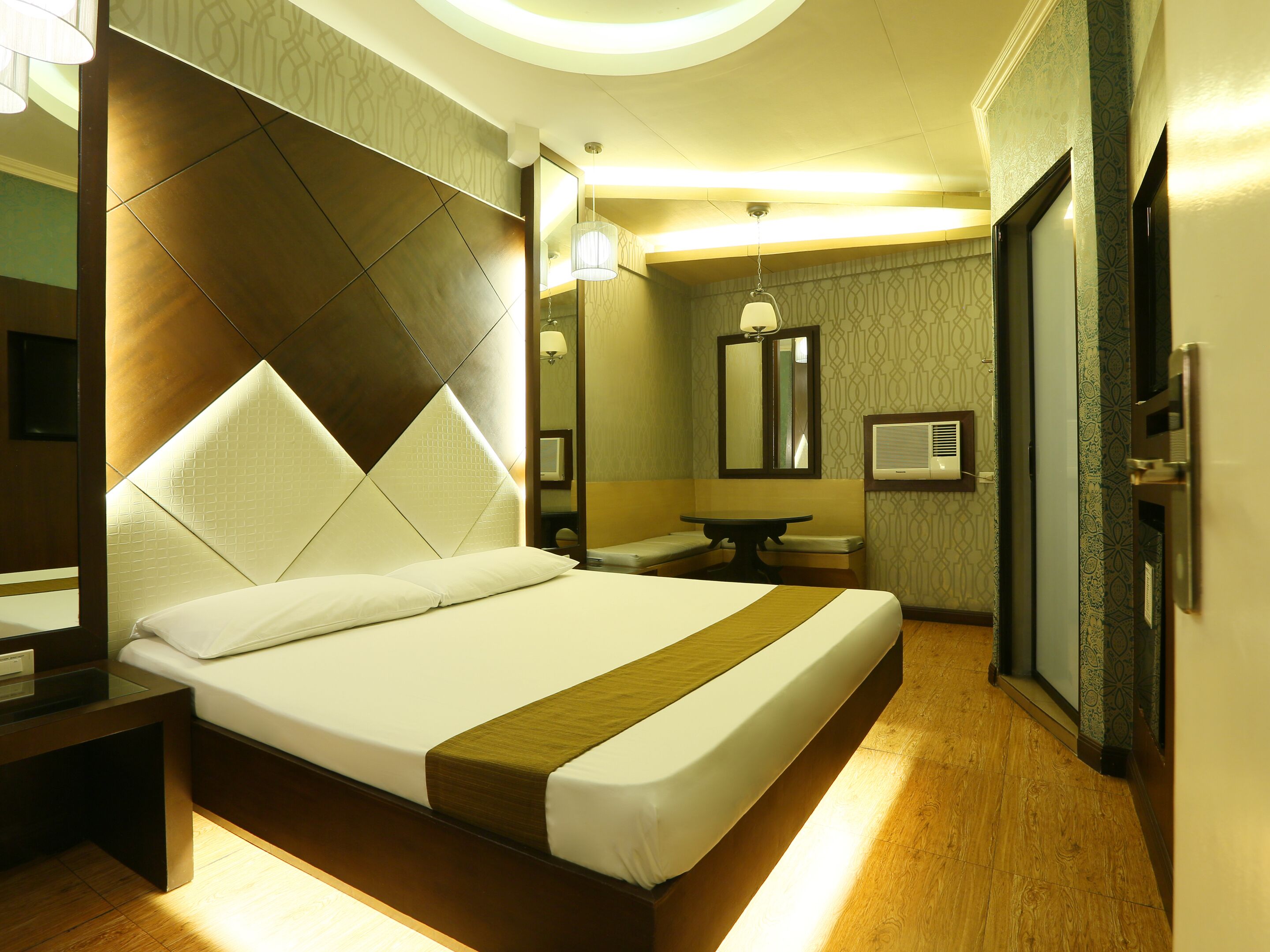 A hotel room with a large bed, a bedside table, a mirror, a small table with a chair, and a wall-mounted air conditioner.