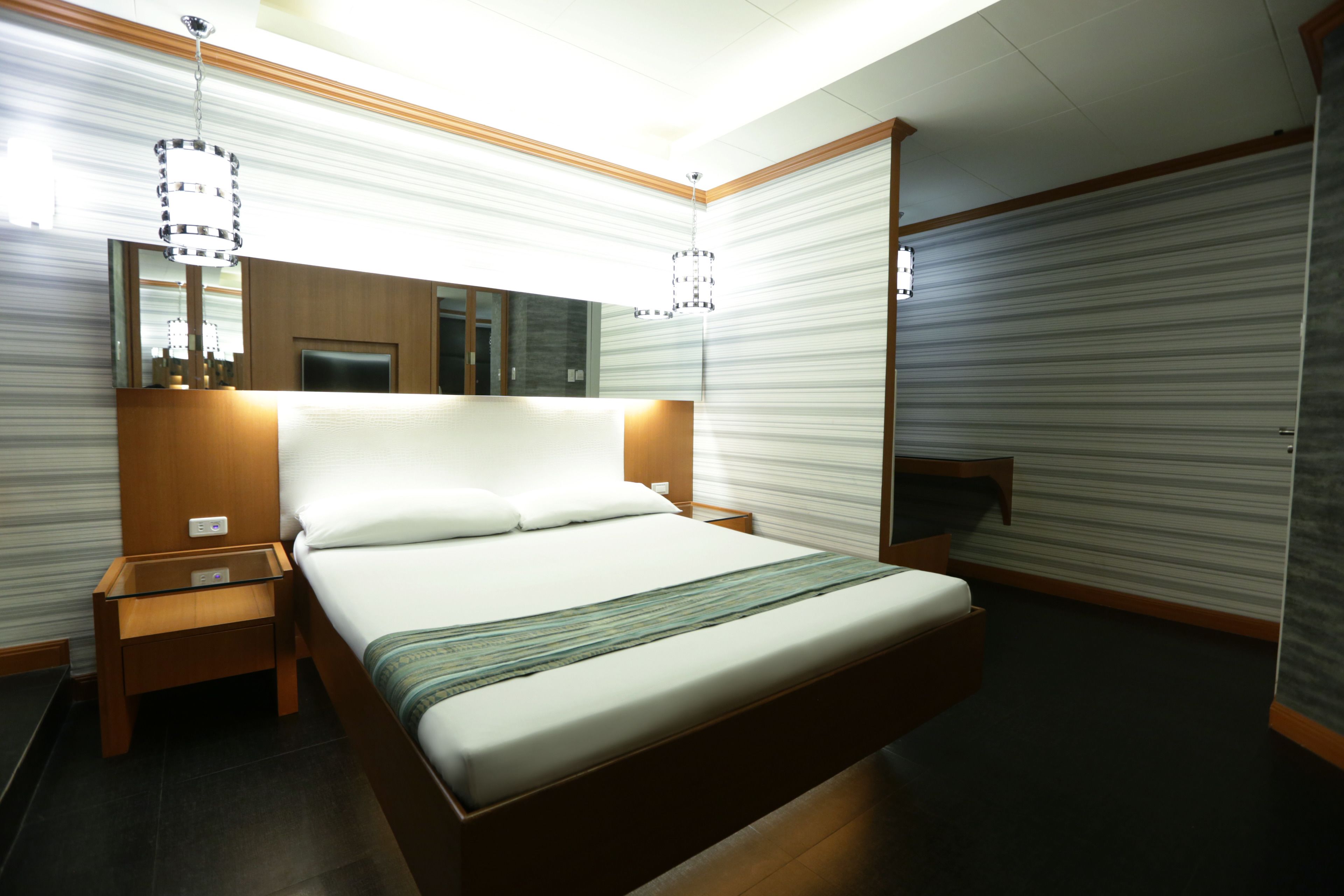 A modern hotel room with a large bed, wooden headboard, bedside tables, a mirror, and a chandelier.