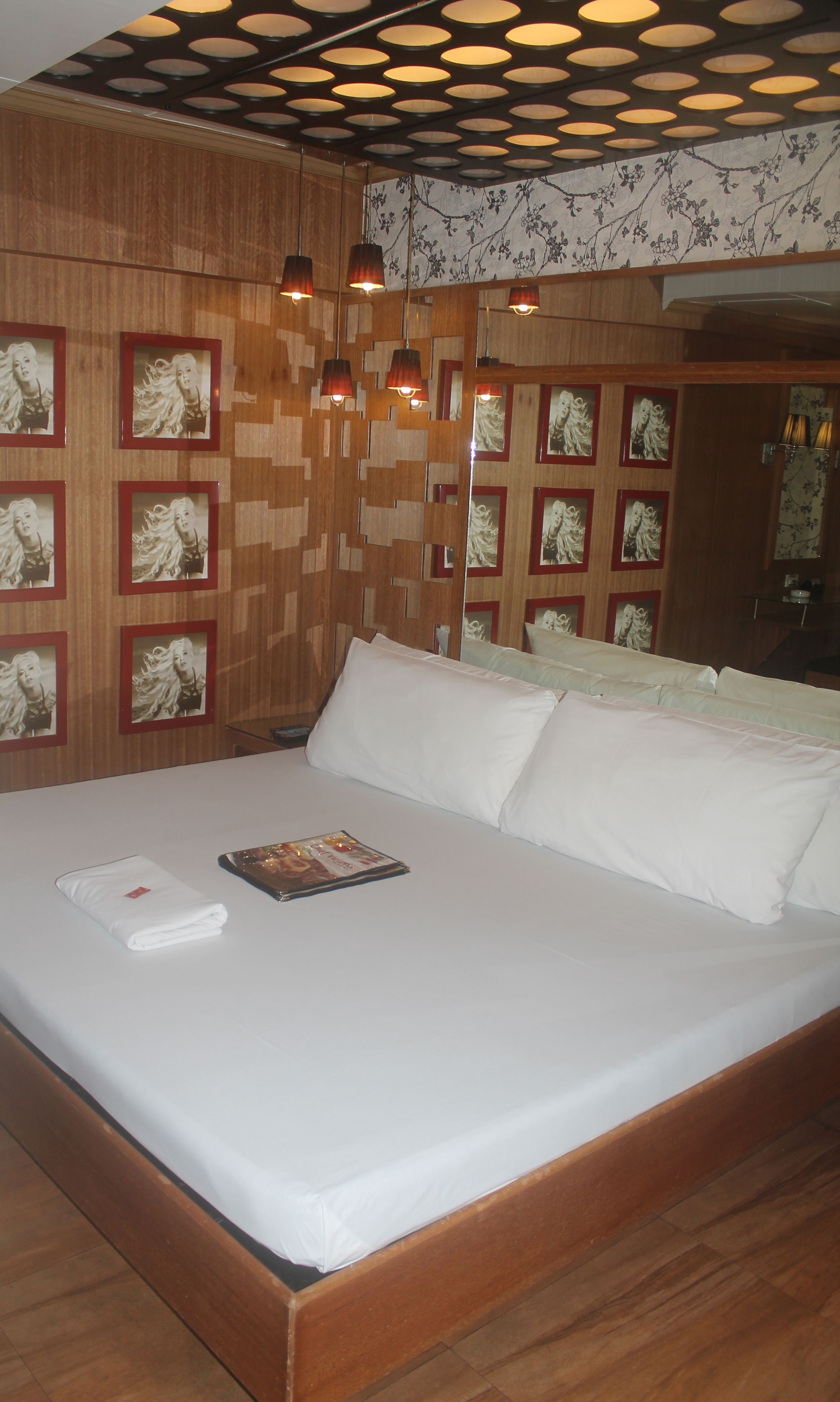 A neatly made bed with white linens, a magazine on the bed, and a folded towel on the side. The room has wooden paneling on the walls and a patterned wallpaper.