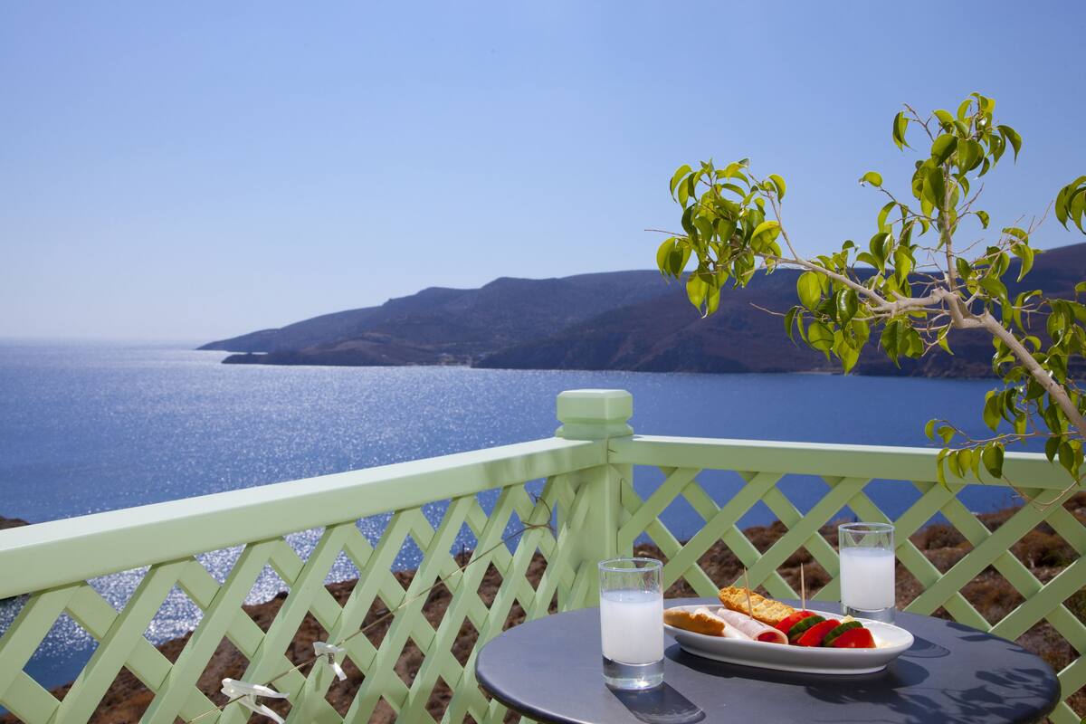 Une table avec une assiette de nourriture et deux verres contenant une boisson blanche, le tout placé devant un décor composé d’une mer bleue et dégagée et de collines lointaines.