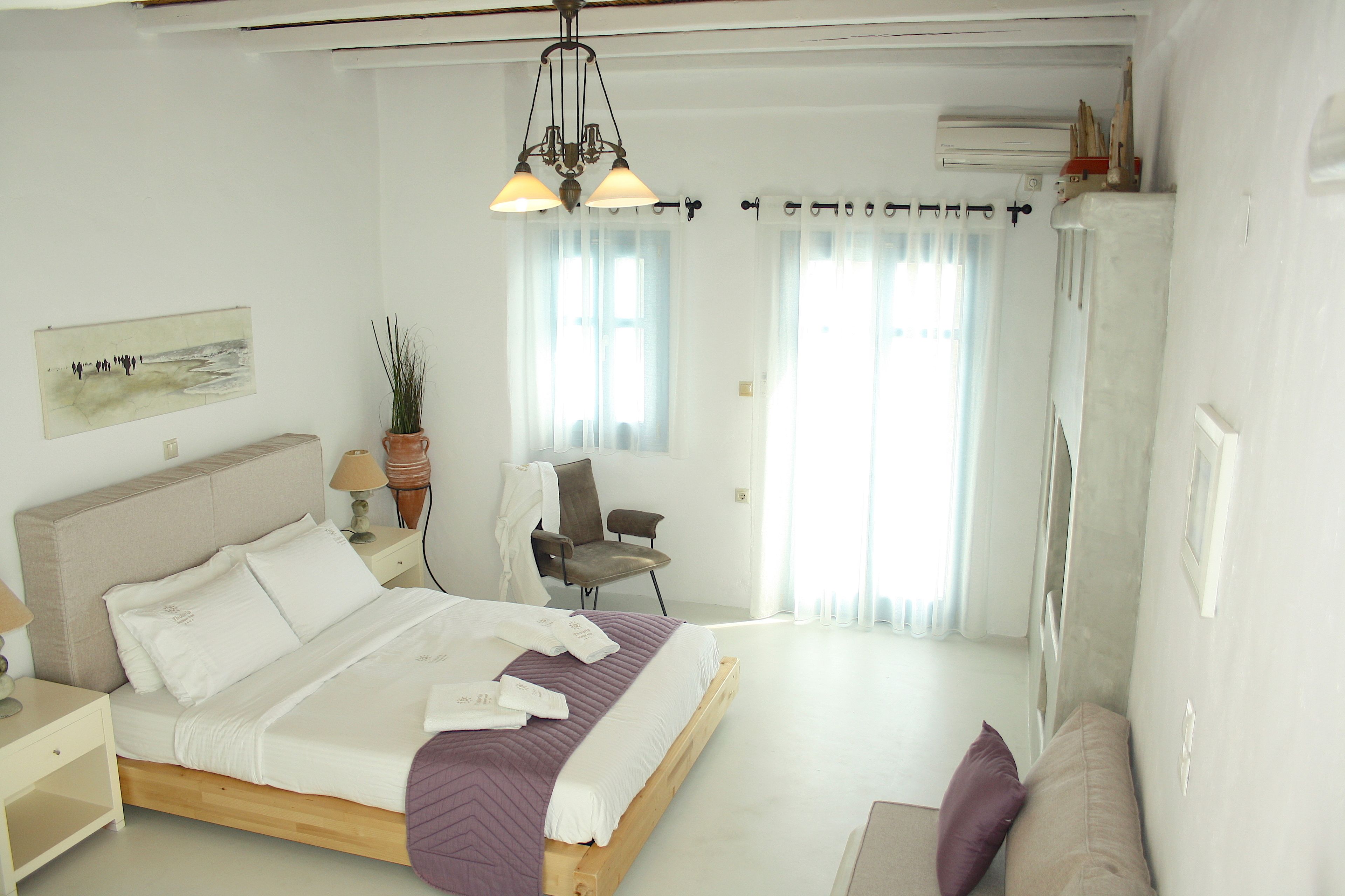Un dormitorio con cama, una silla, una mesita de noche, una lámpara y un ventanal con cortinas.