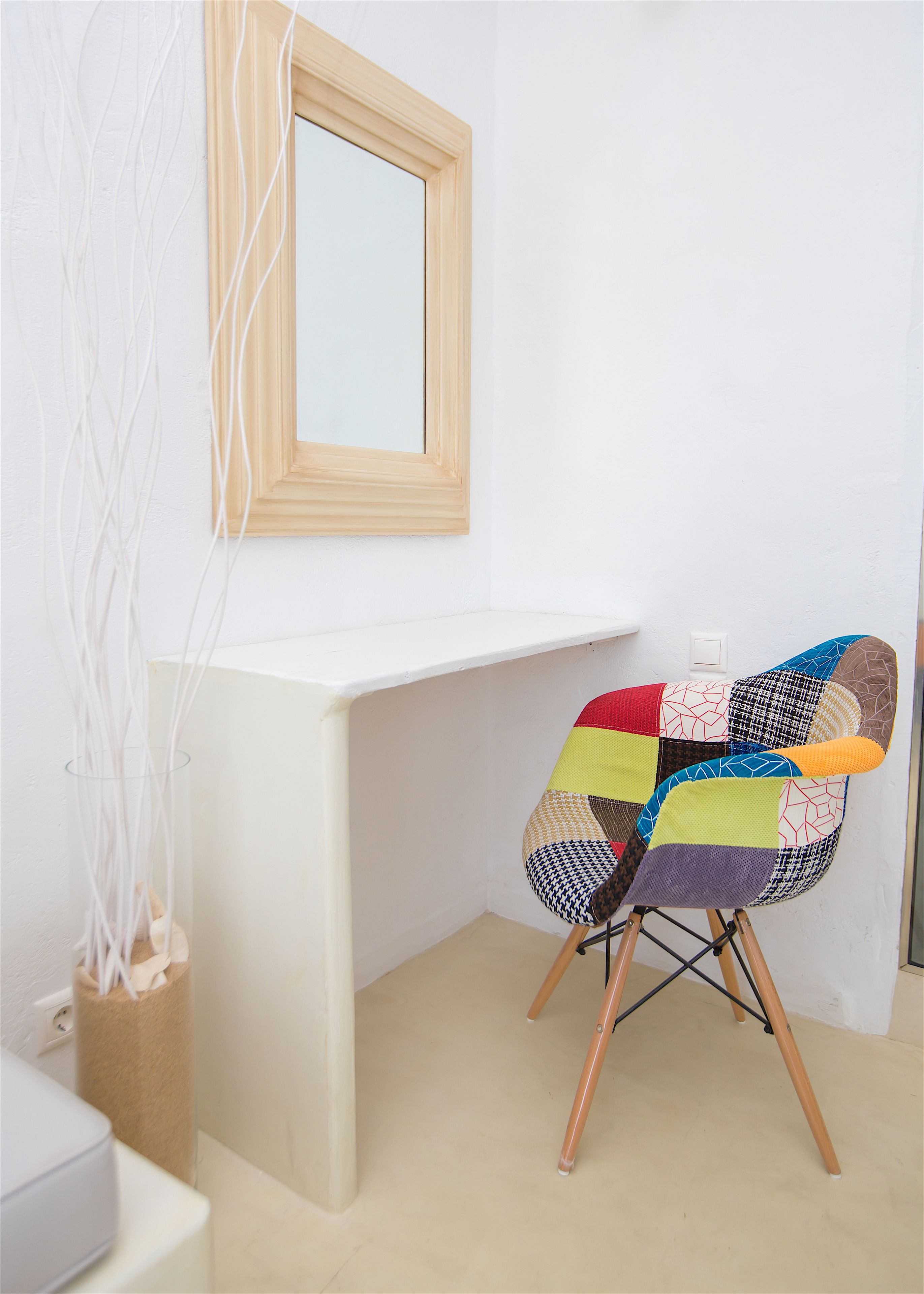 A corner of a room with a white desk, a colorful chair, a mirror, and a vase with white branches.