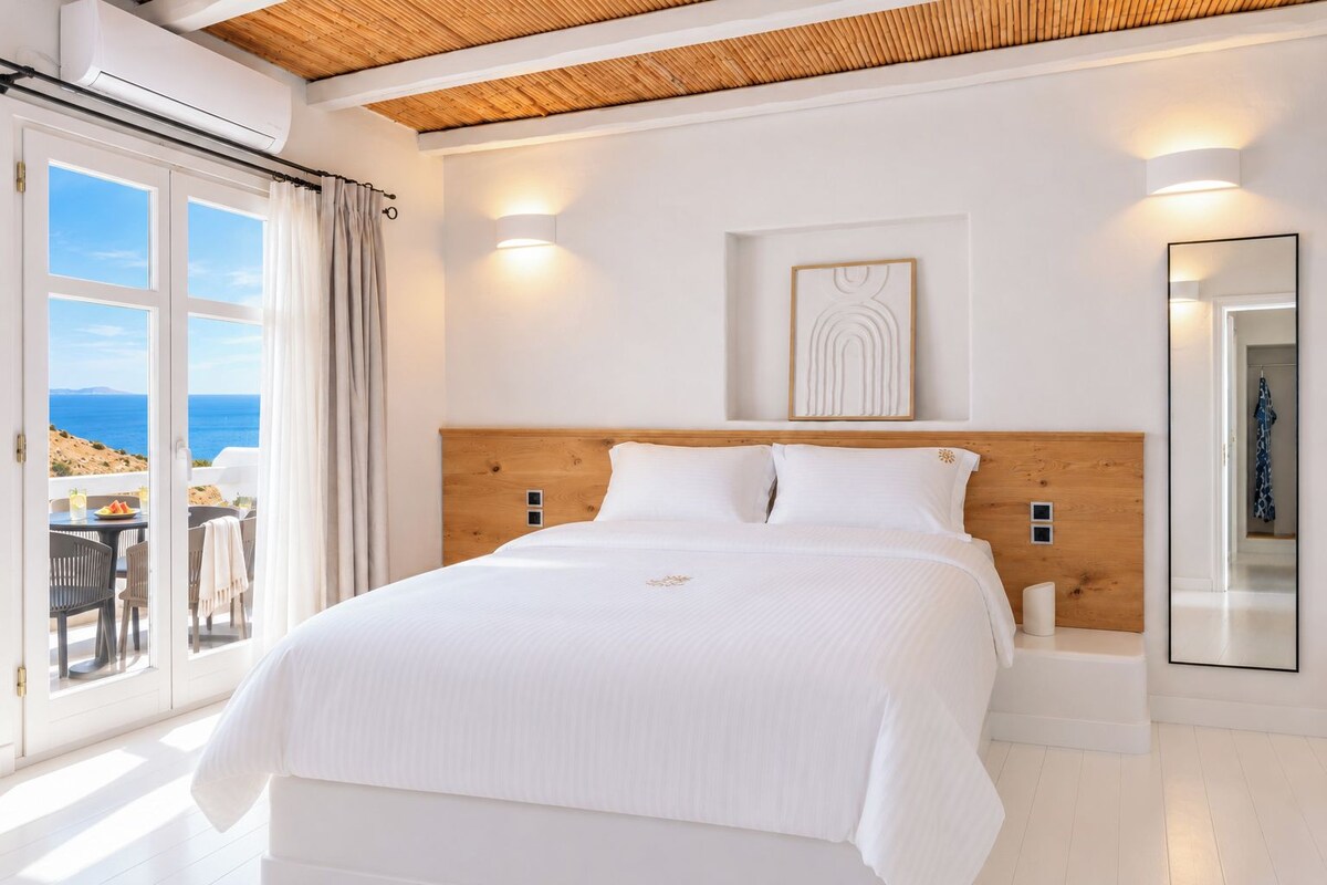 Un dormitorio con una cama grande, cabecera de madera, vista al mar y un balcón con mobiliario exterior.