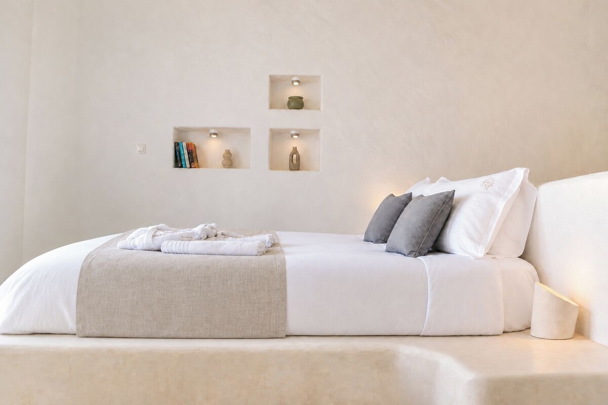 Una cama bien hecha con una manta color beige y almohadas, una mesita de noche con una lámpara y estantes en la pared con objetos decorativos.