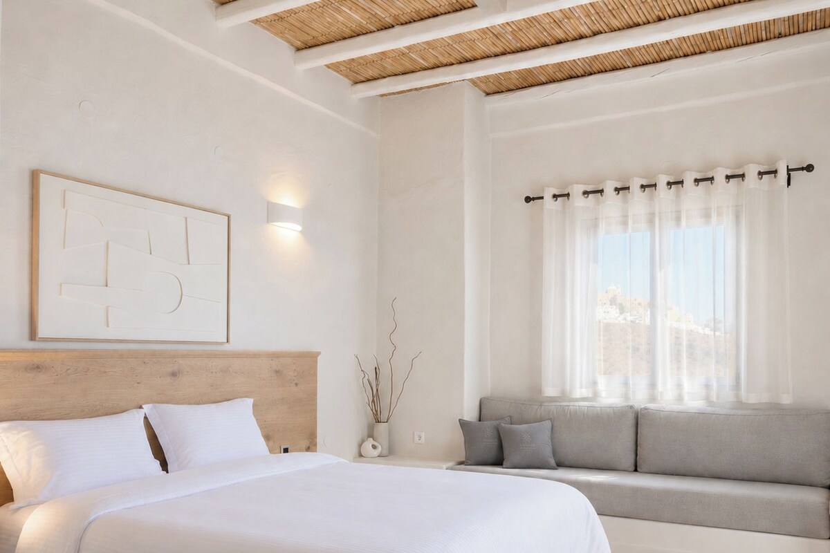Un dormitorio con cama, sofá, una obra de arte en la pared y ventana con cortinas translúcidas.