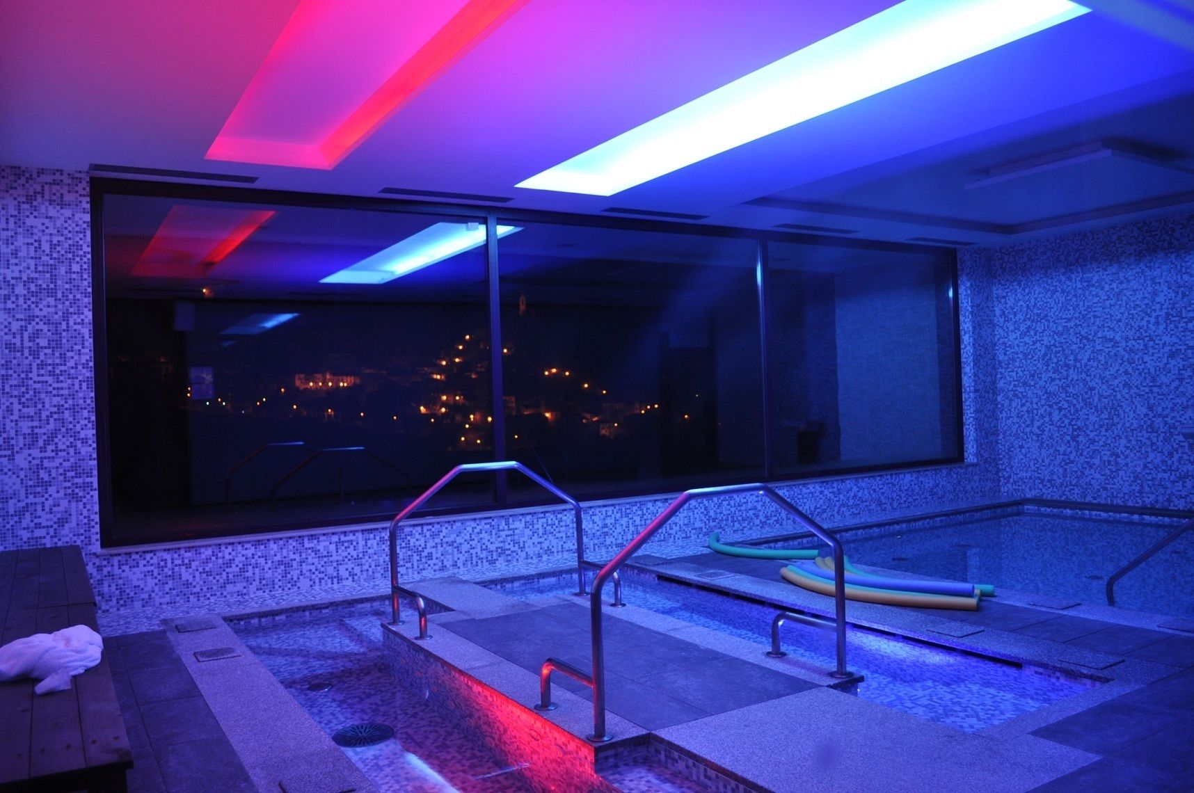 Un espace piscine intérieur moderne, doté d’une grande fenêtre, de rampes en métal et offrant une vue sur le paysage urbain illuminé la nuit.