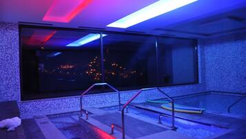 Un espace piscine intérieur moderne, doté d’une grande fenêtre, de rampes en métal et offrant une vue sur le paysage urbain illuminé la nuit.