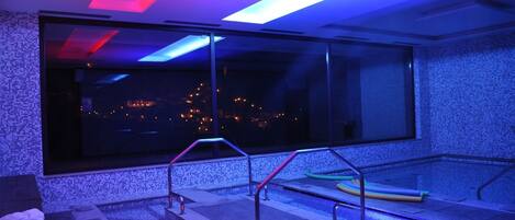 Un área de piscina interior moderna con un amplio ventanal, barandales de metal y vistas a un paisaje urbano nocturno.