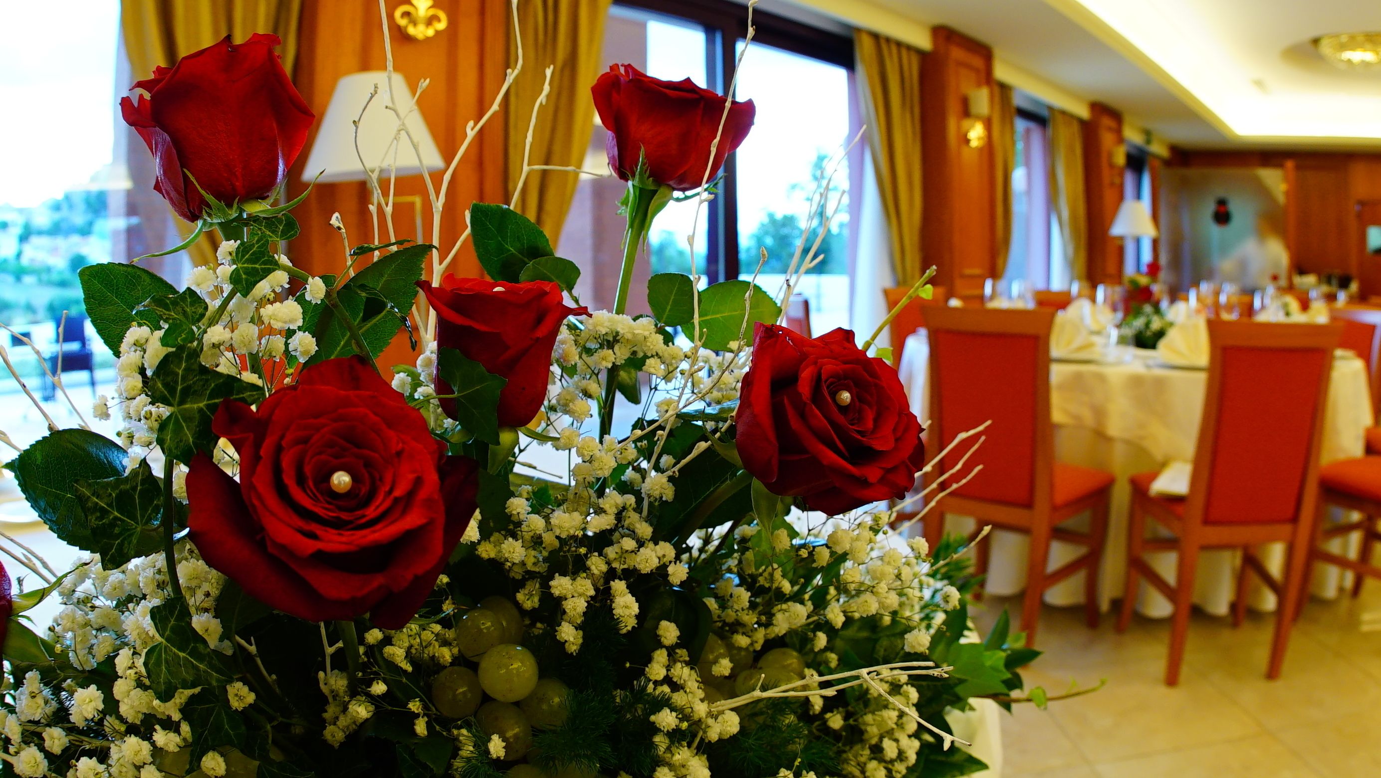A dining area with tables set for a meal, a bouquet of red roses, and a window with curtains.