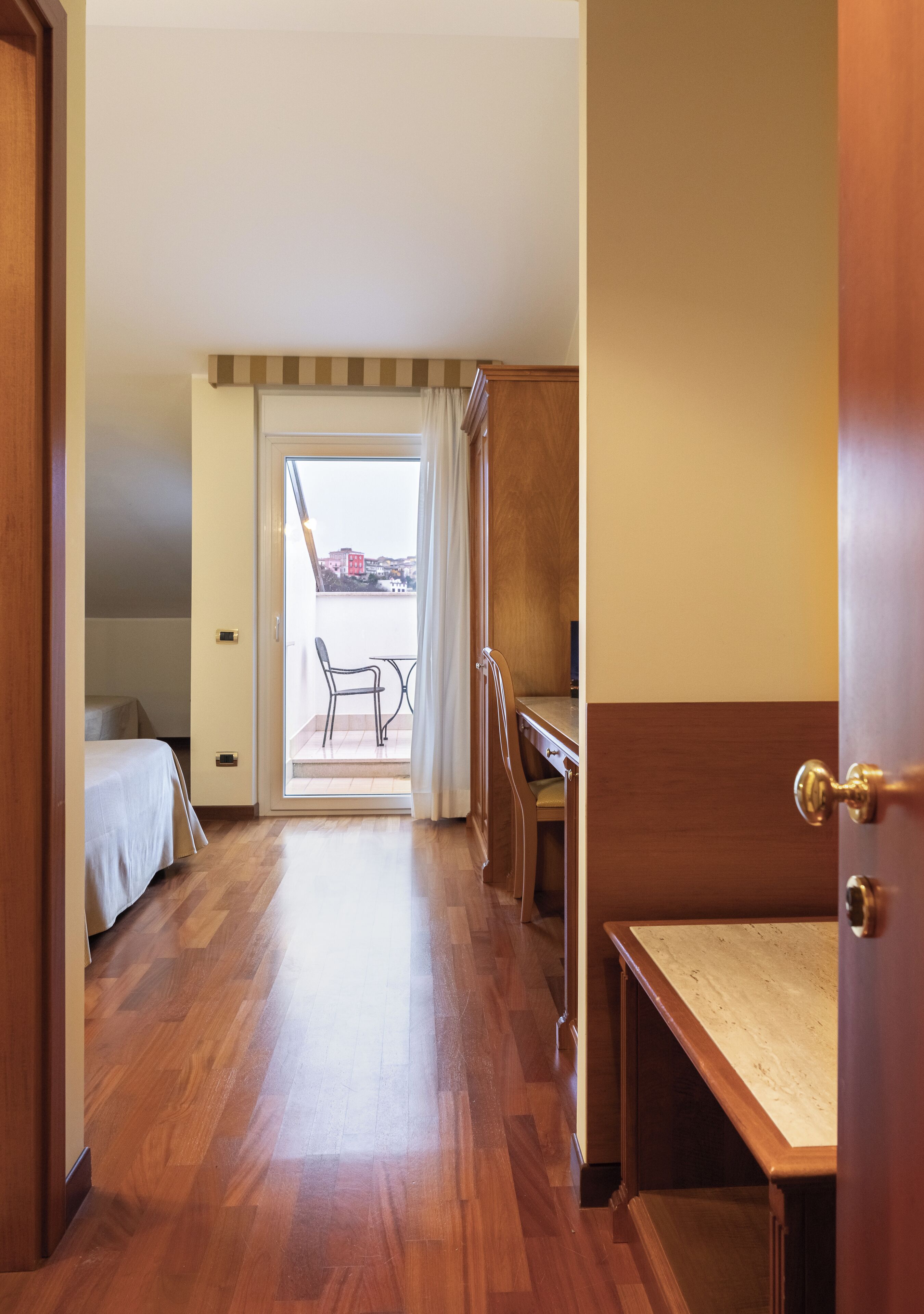 A hotel room with a wooden floor, a bed, a desk, and a chair. There is a sliding door leading to a balcony with a view of buildings.
