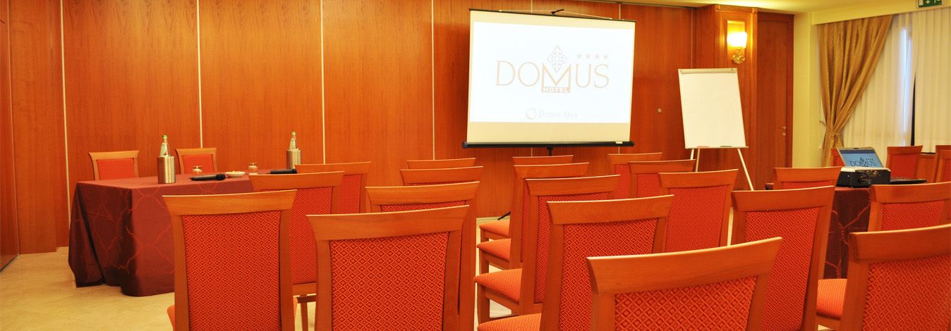 A conference room with rows of wooden chairs, a projection screen, a whiteboard, and a laptop on a table.