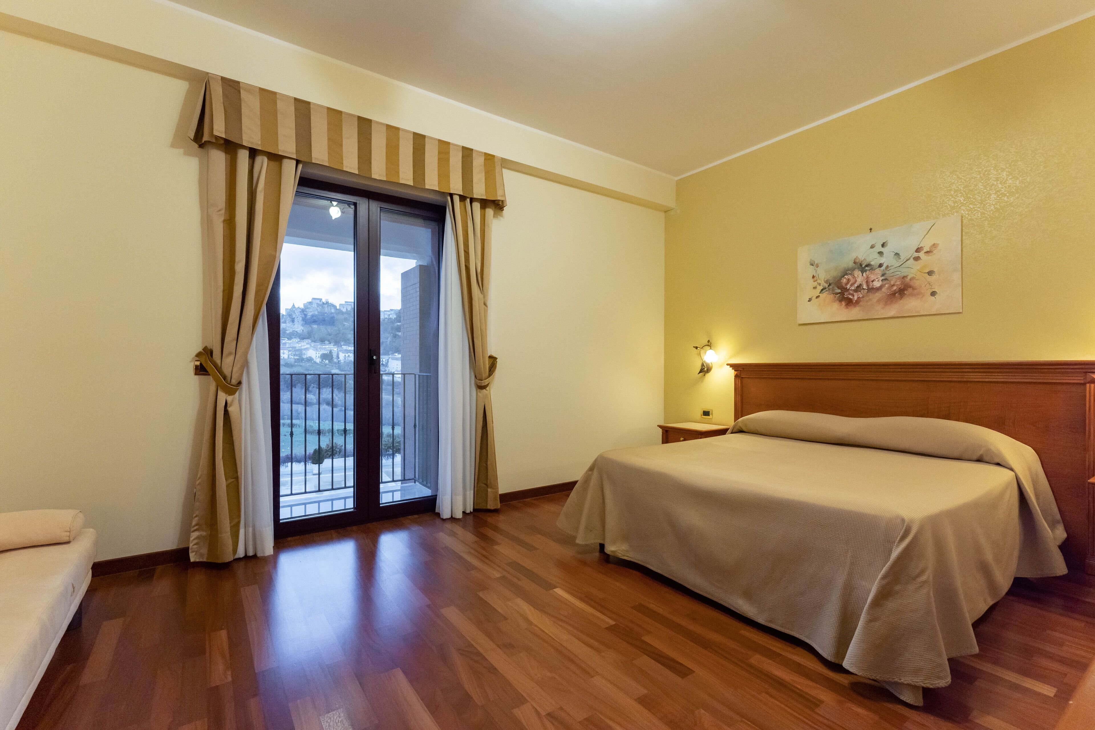 A hotel room with a bed, a nightstand, a painting on the wall, and a view of the outside through a window with curtains.