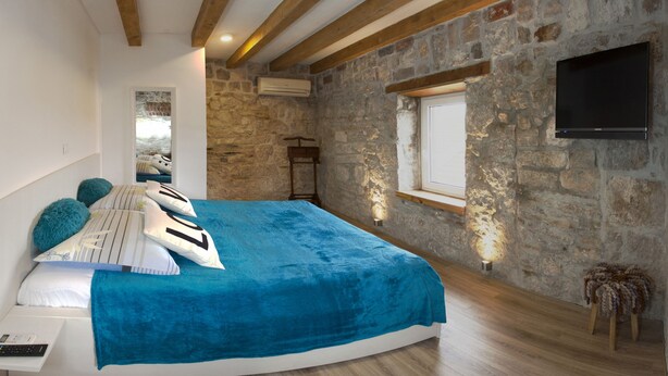 Ein Schlafzimmer mit einer Steinswand, einem Bett mit blauer Decke, einem an der Wand befestigten Fernseher und einer Holzbalkendecke.