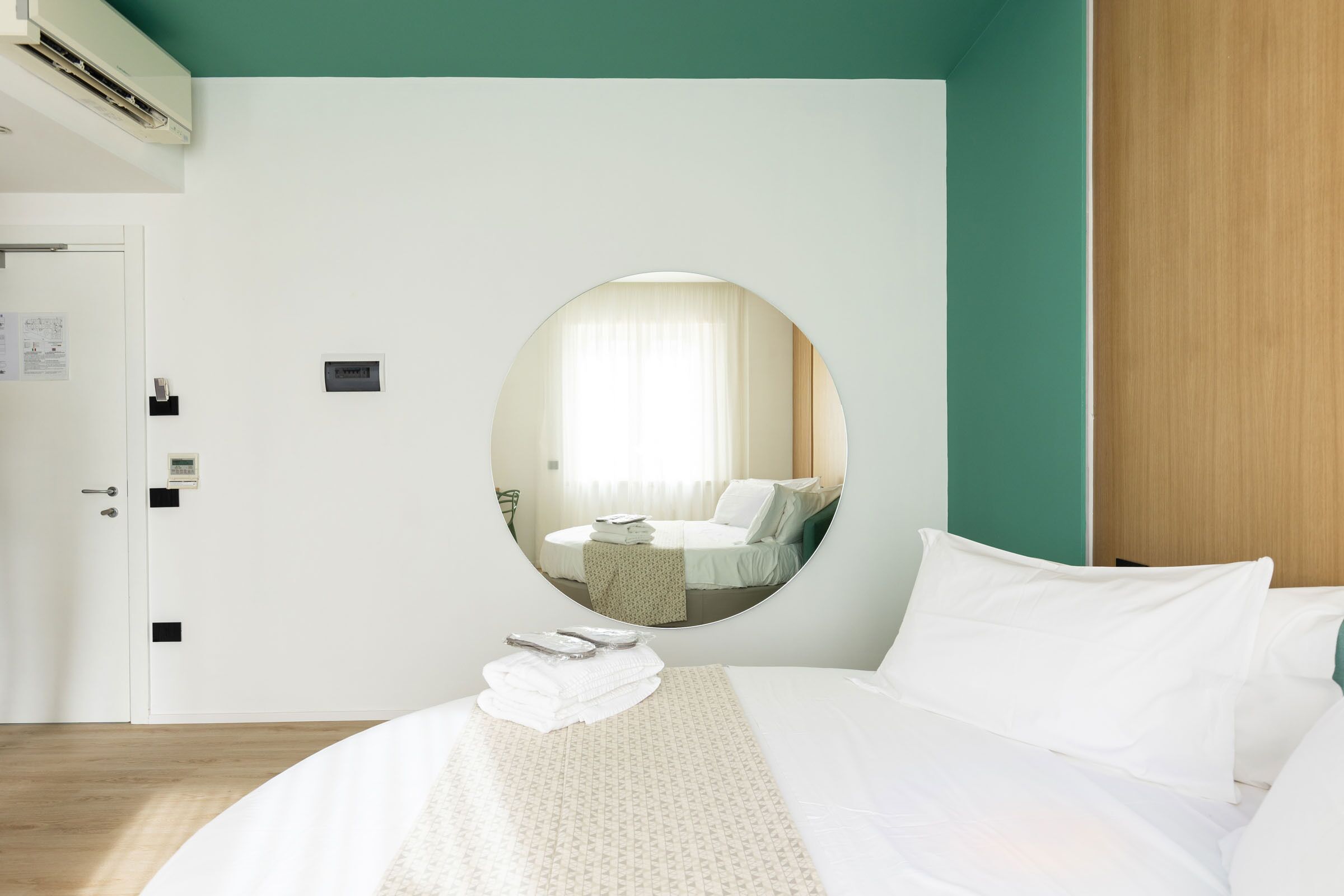Habitación de hotel con cama, espejo redondo, puerta y aire acondicionado instalado en la pared.