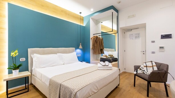 A modern hotel room with a large bed, a bedside table with a plant, a chair, and a coat rack with clothes. - Hotel Plana (Caserta)