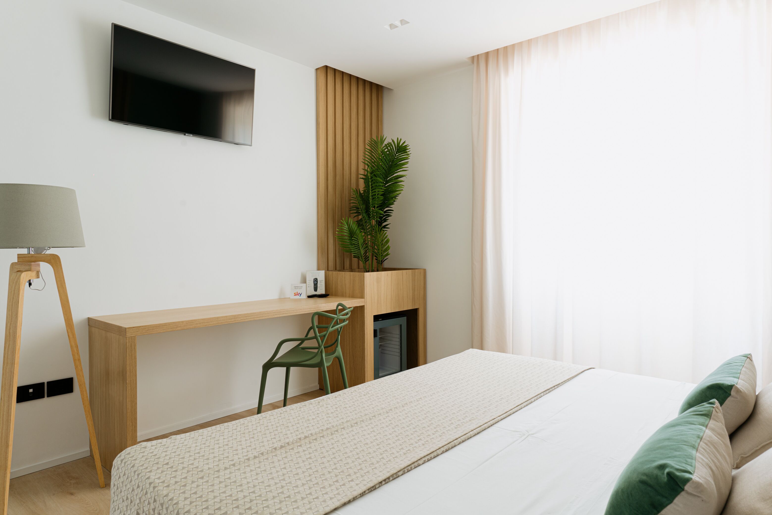 A modern hotel room with a bed, a desk, a chair, a TV, and a plant.