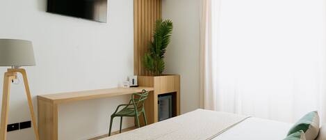 A modern hotel room with a bed, a desk, a chair, a TV, and a plant.