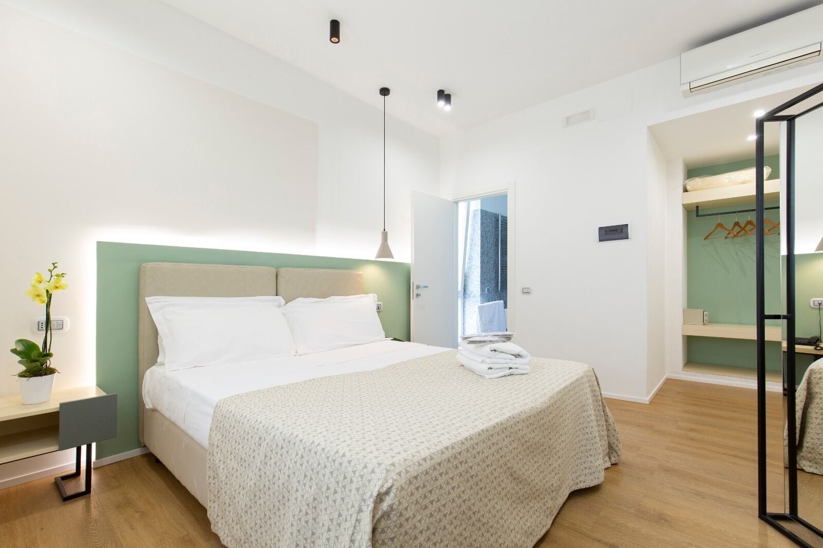 Una habitación de hotel moderna con una cama, una mesita de noche, una planta en maceta y vistas de la ciudad a través de una puerta abierta.