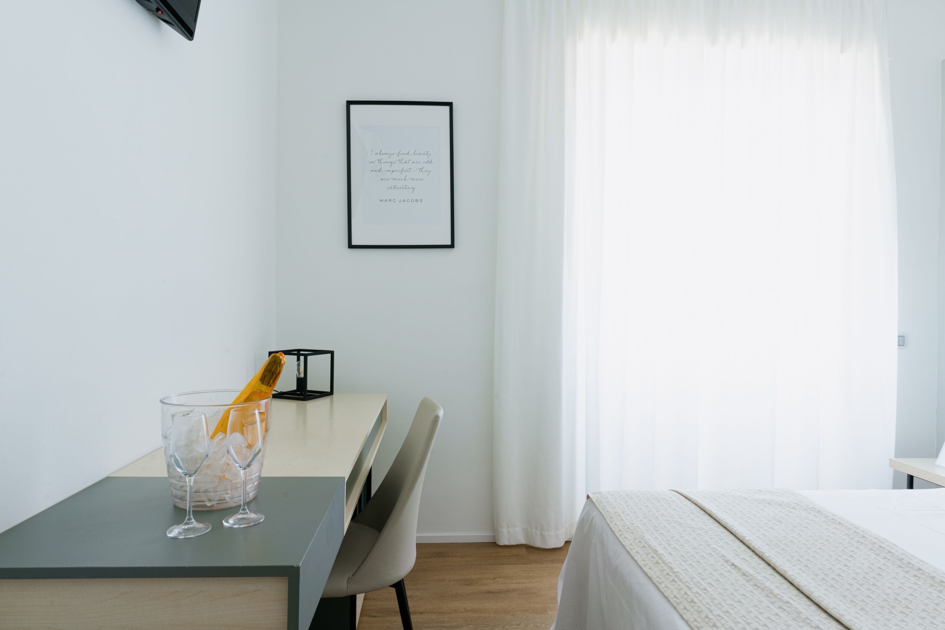 A minimalist hotel room with a bed, a desk with a glass of champagne, a framed quote on the wall, and sheer curtains.