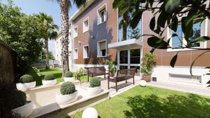 A modern building with a flat roof, large windows, and a well-maintained garden with benches and potted plants. - Hotel Plana (Caserta)