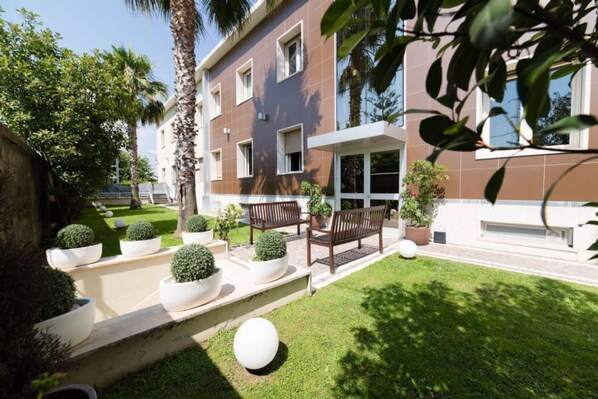A modern building with a flat roof, large windows, and a well-maintained garden with benches and potted plants. - Hotel Plana (Caserta)