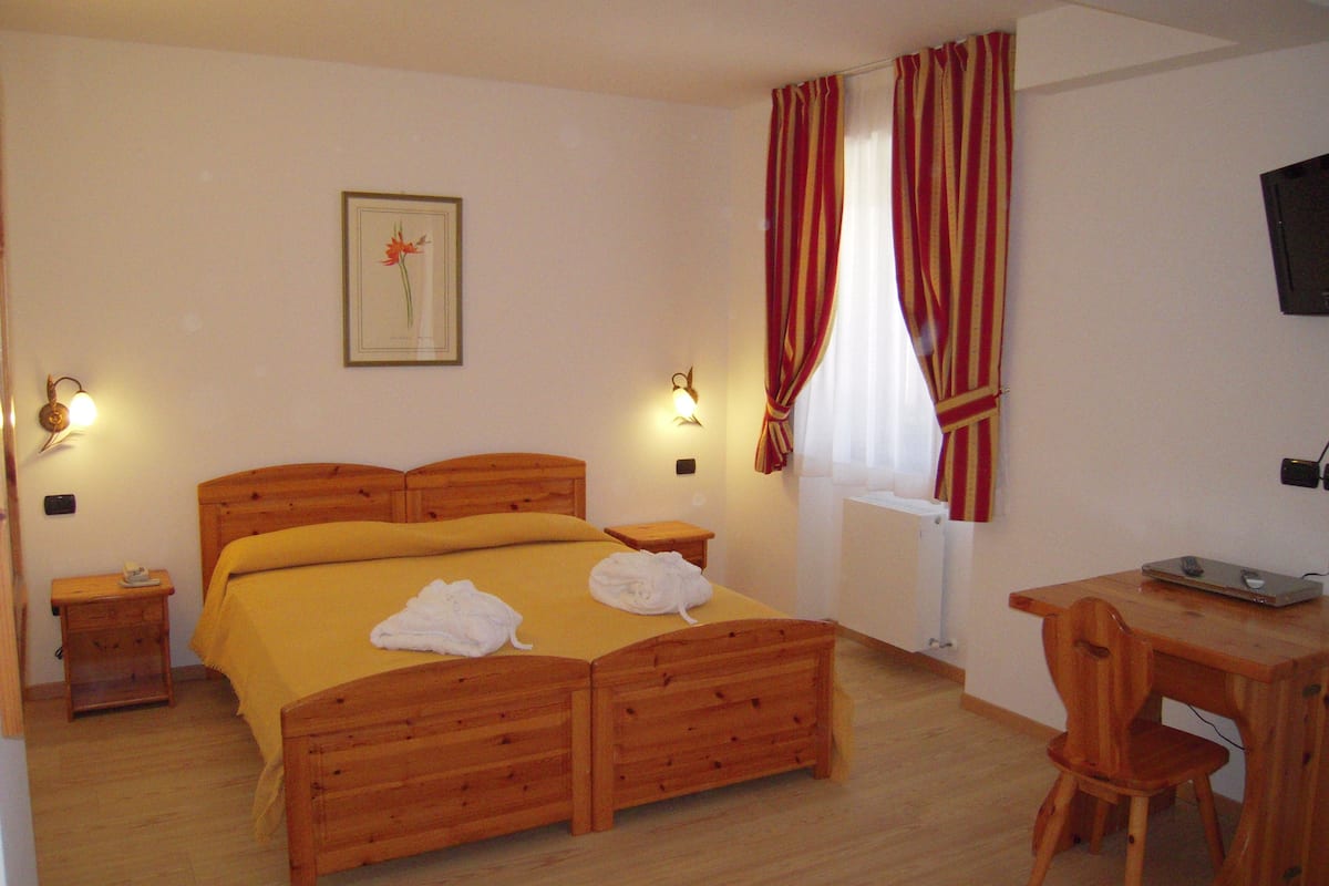 A hotel room with a wooden bed, two bedside tables, a desk with a chair, a television, and a window with curtains.