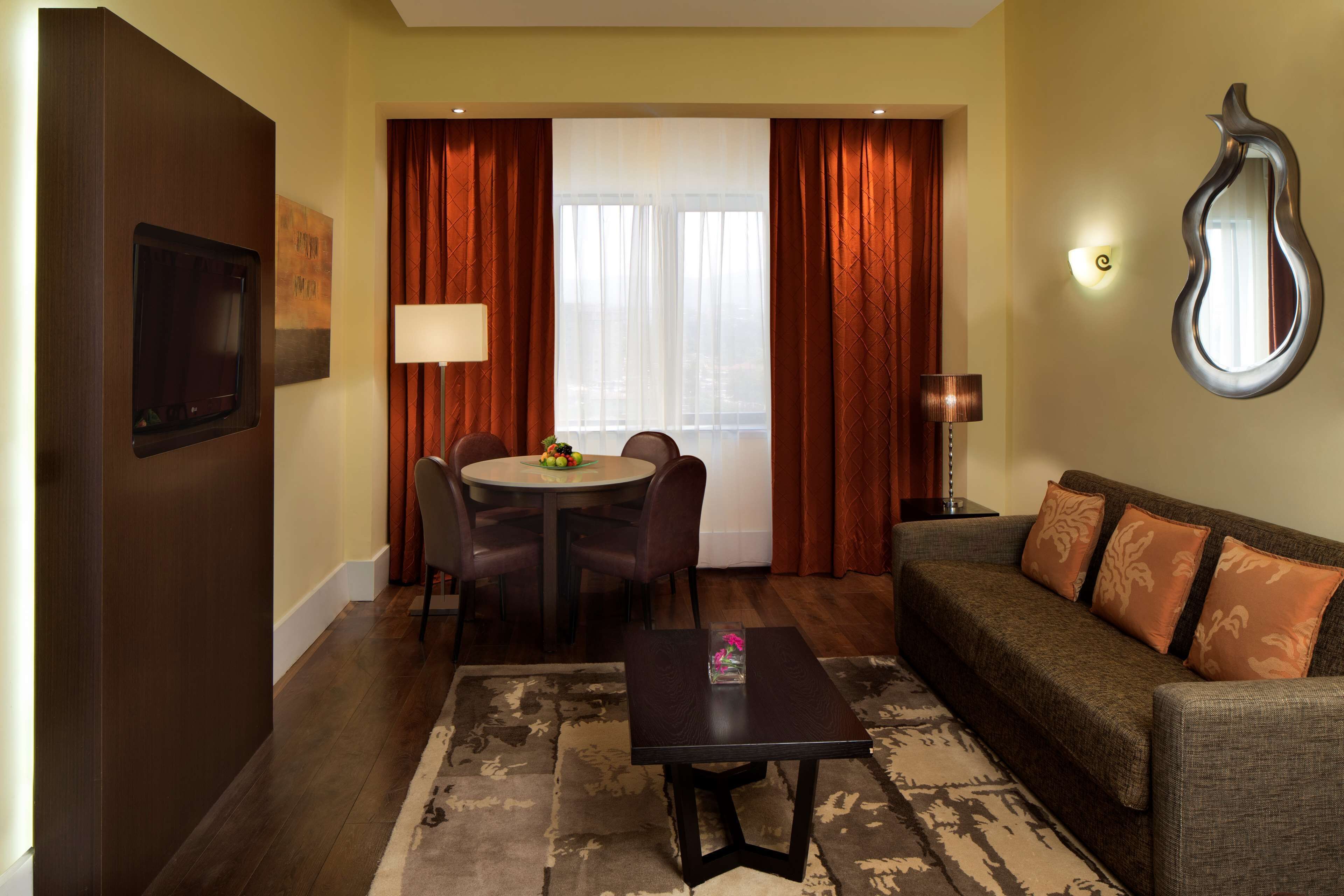 Habitación de hotel con comedor, sofá, televisor y un amplio ventanal con cortinas.
