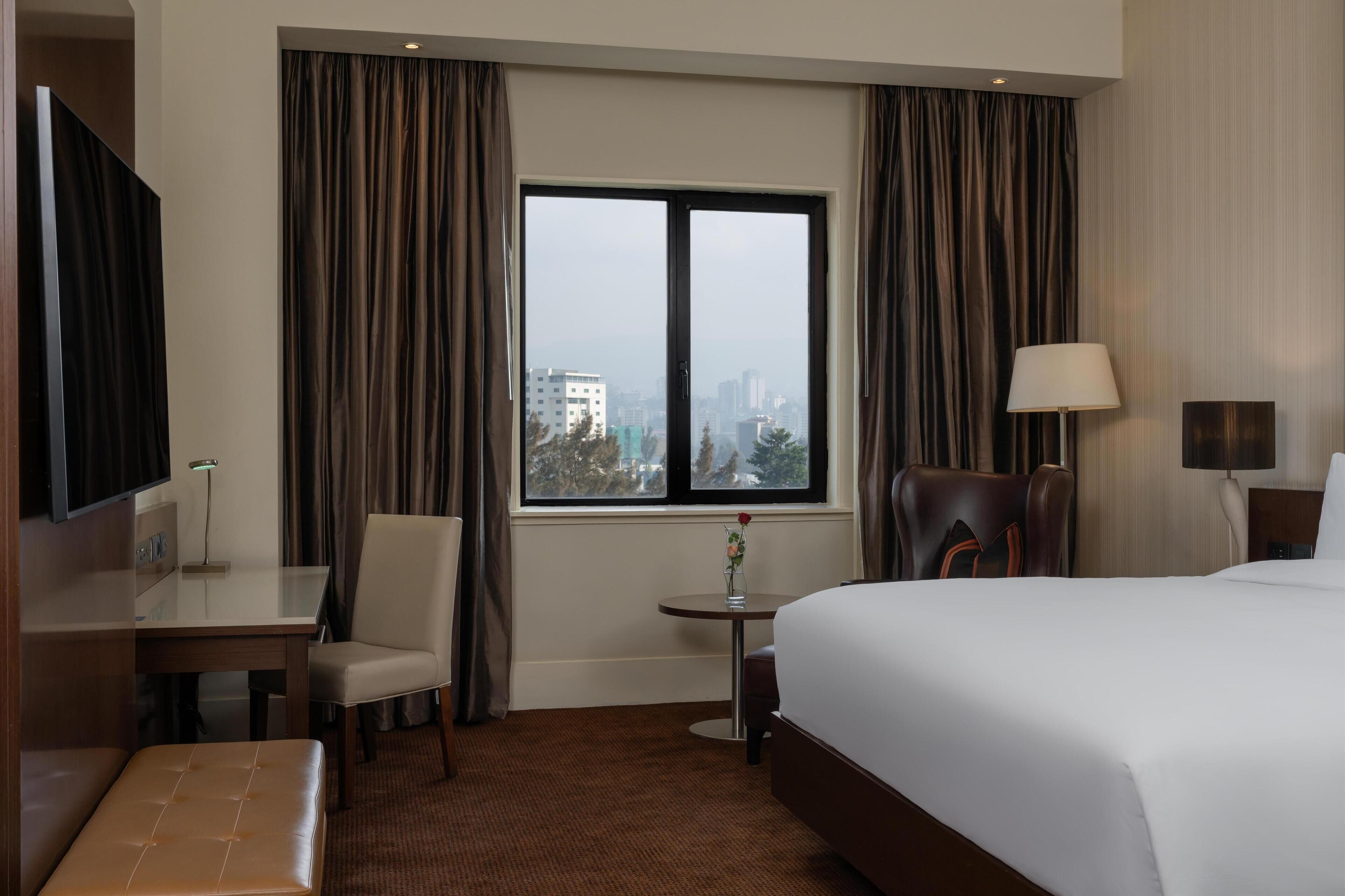 A hotel room with a bed, a desk, a chair, a small table with a vase, and a view of a cityscape through a window.