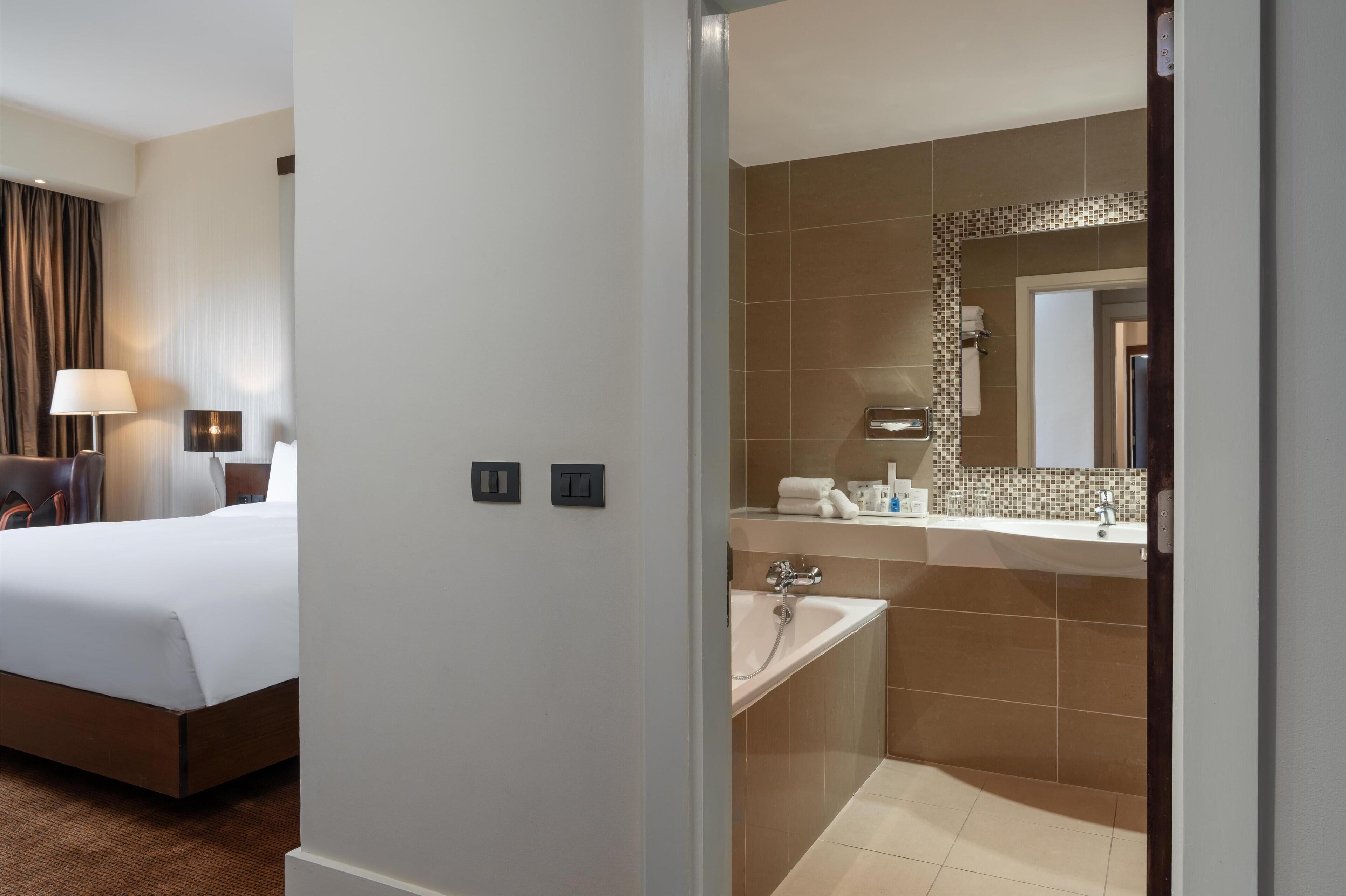 A hotel room with a bed, bedside table, lamp, and a bathroom with a sink, mirror, and shower.