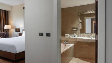A hotel room with a bed, bedside table, lamp, and a bathroom with a sink, mirror, and shower.