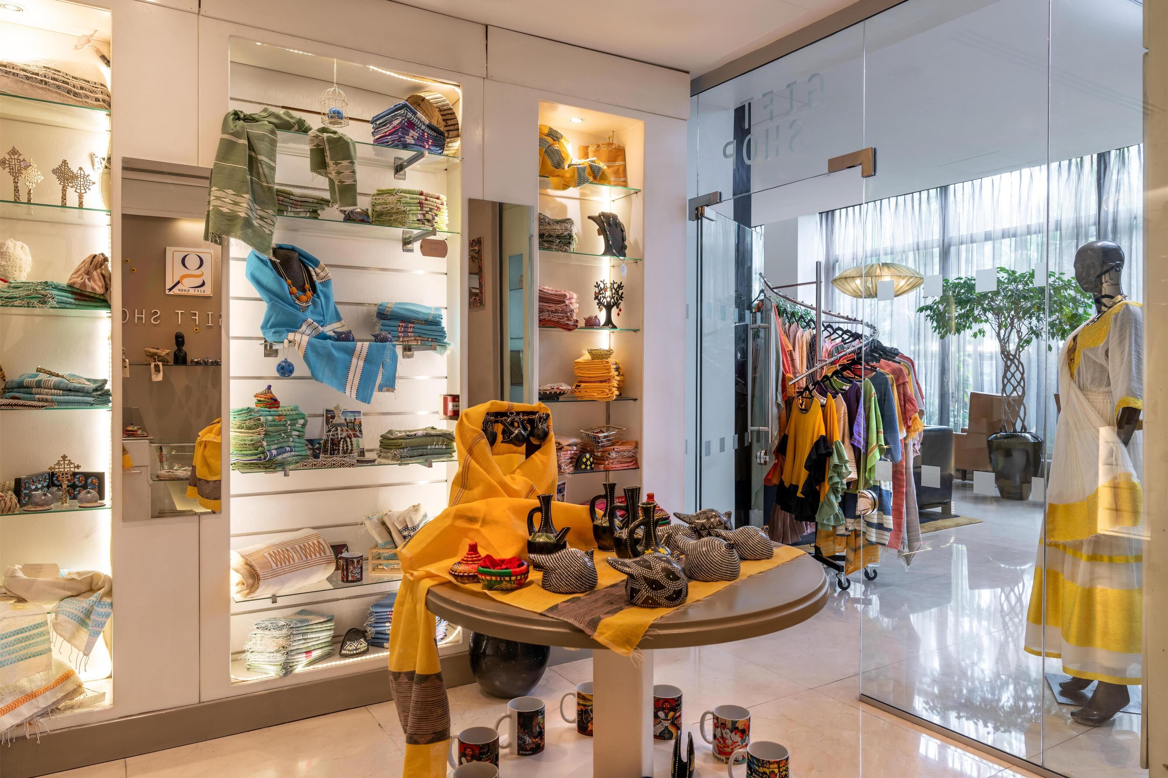 Una boutique bien iluminada con textiles y accesorios ordenados en estantes y mesas, un maniquí vestido con traje tradicional, y un perchero con diversas prendas de vestir.