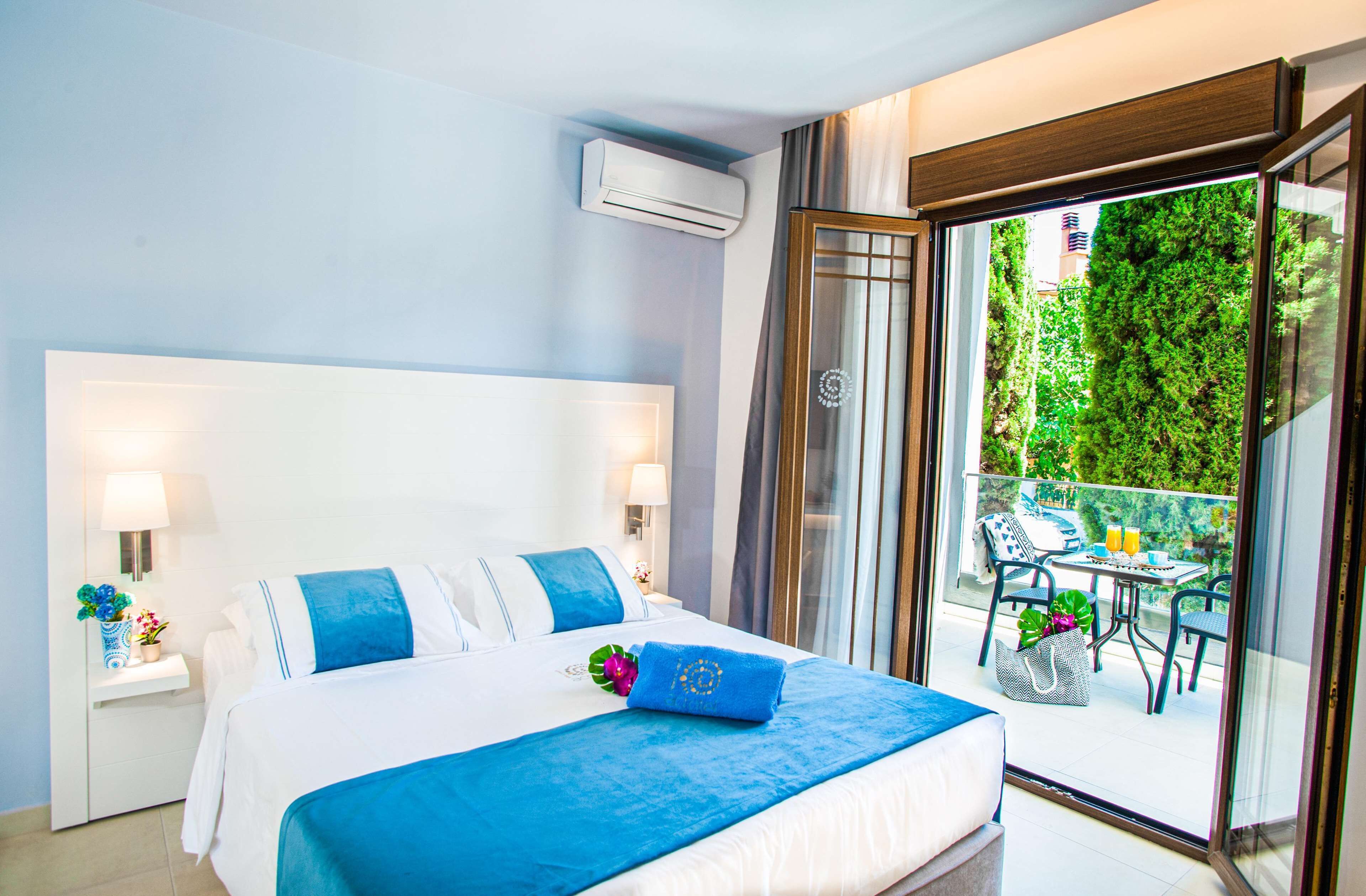 A hotel room with a large bed, a balcony with outdoor seating, and a view of greenery.