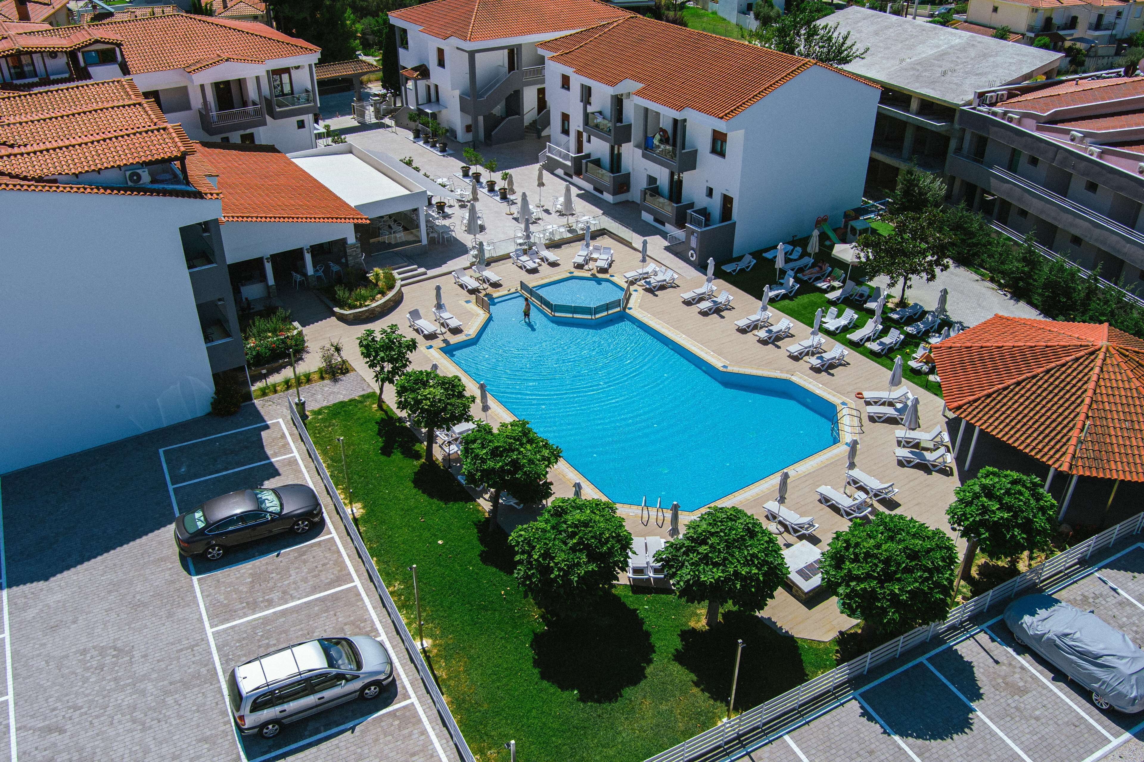 Vista aérea de um hotel com uma piscina central cercada por espreguiçadeiras e guarda-sóis, várias varandas e um estacionamento.