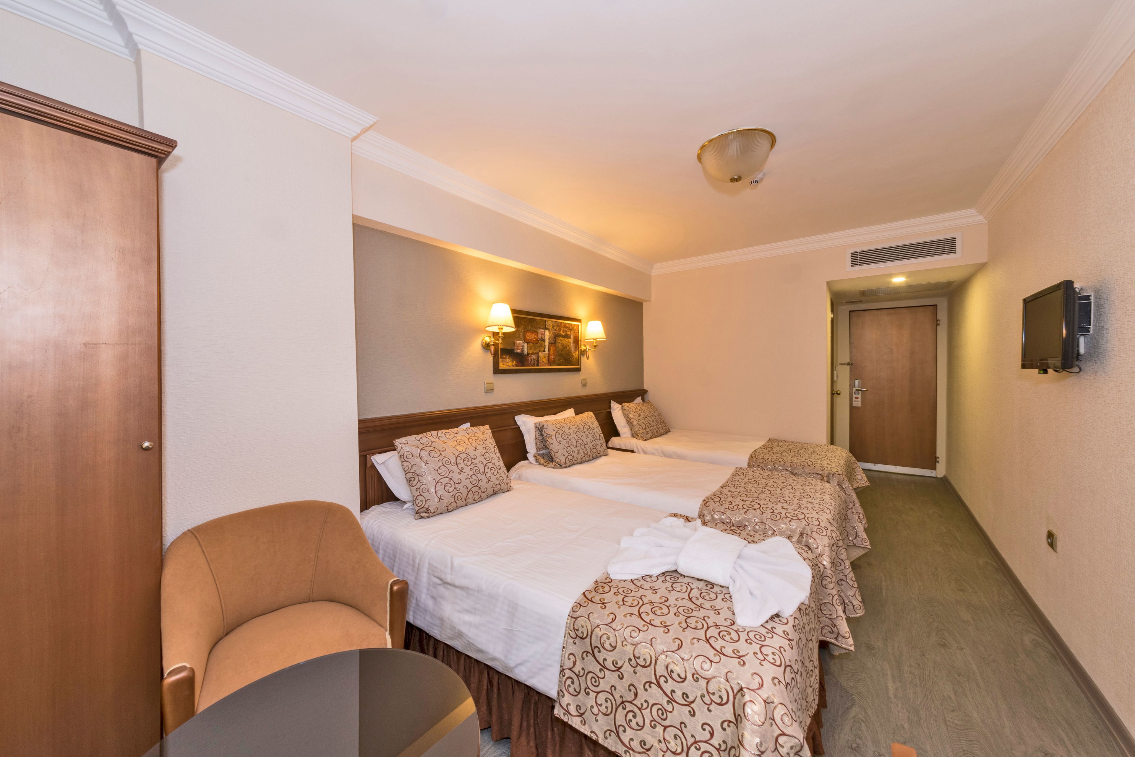 A hotel room with two beds, a wooden wardrobe, a chair, and a television mounted on the wall.