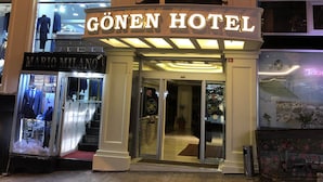 L’entrée de l’hôtel GÖNEN est clairement indiquée par un panneau, et sa devanture, visible, présente une boutique de vêtements. Le sol est pavé de briques.