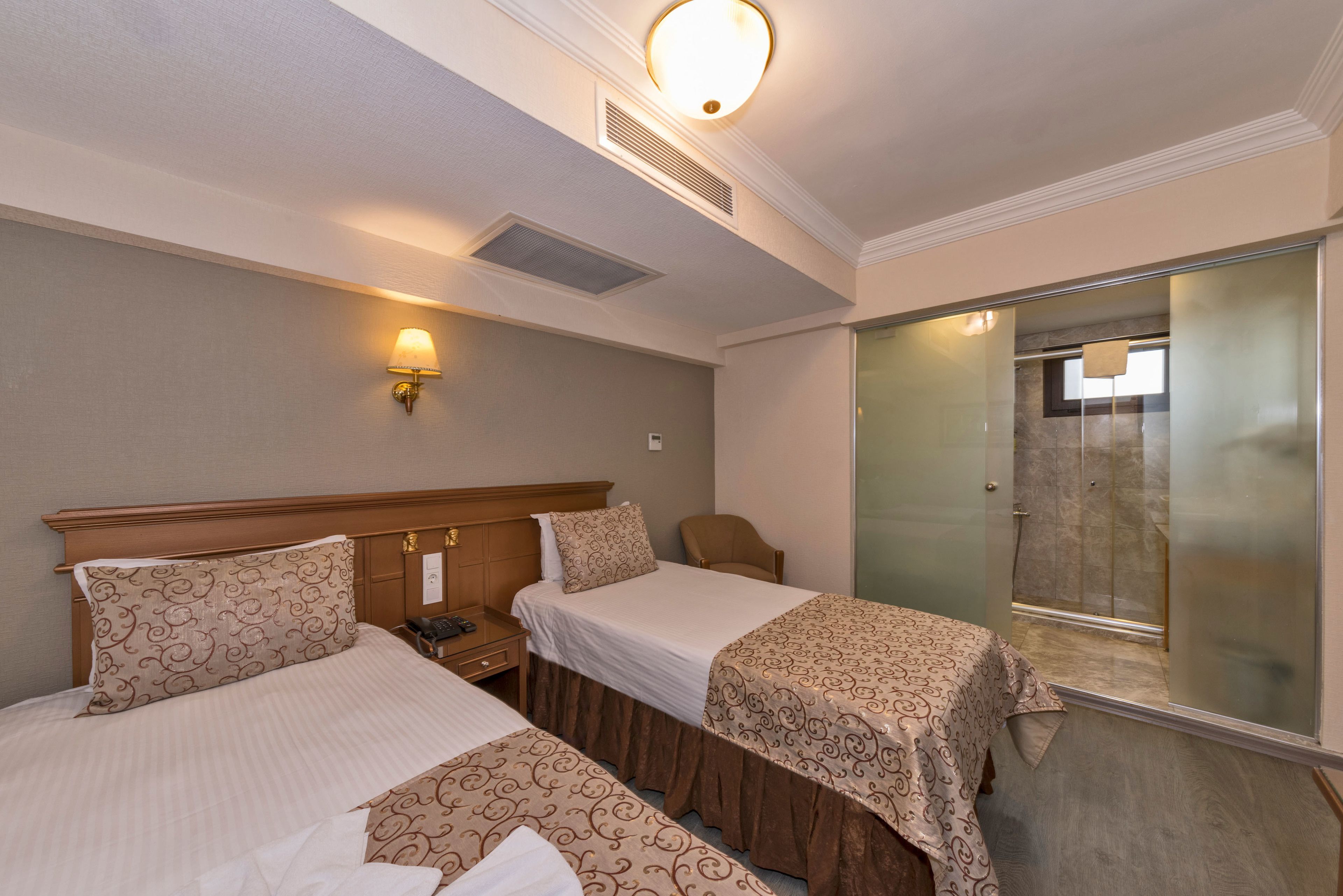 Habitación de hotel con dos camas, una mesita de noche, una silla, cabina de ducha de vidrio y una lámpara fijada a la pared.