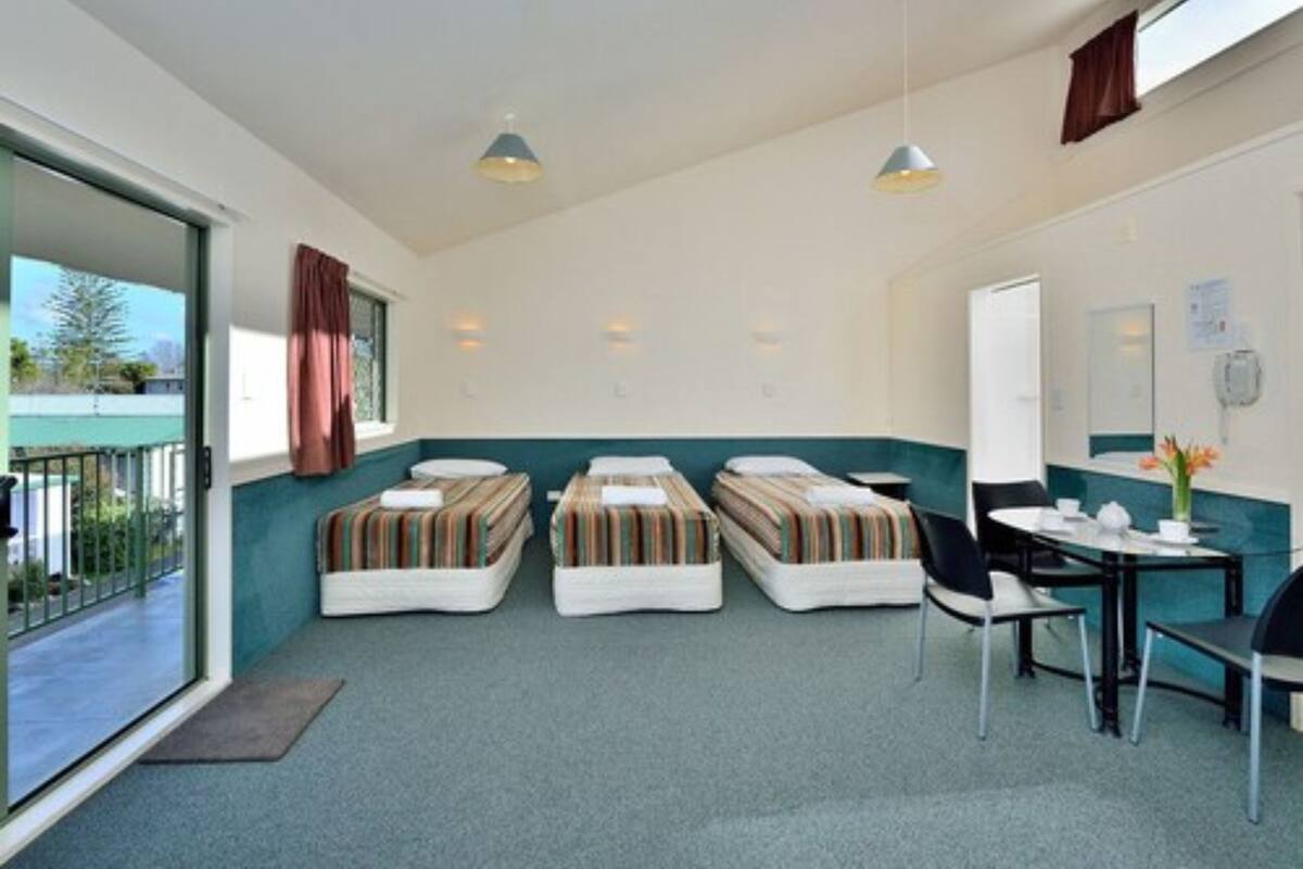 Habitación de hotel con tres camas individuales, una mesita con sillas, una ventana con cortinas y una puerta.
