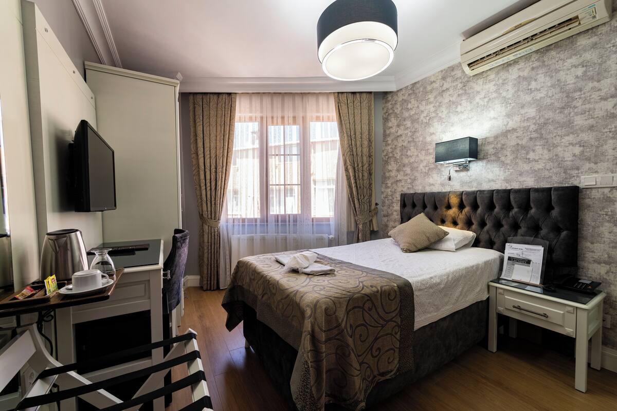 Habitación de hotel con cama, mesita de noche, televisor de pantalla plana montado en la pared, ventana con cortinas e iluminación en el techo.