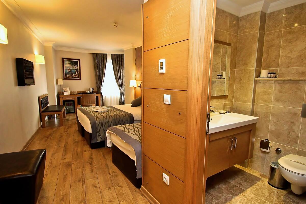 Habitación de hotel con armario de madera, cama, escritorio con silla, mesita de noche con lámpara y baño con lavamanos e inodoro.
