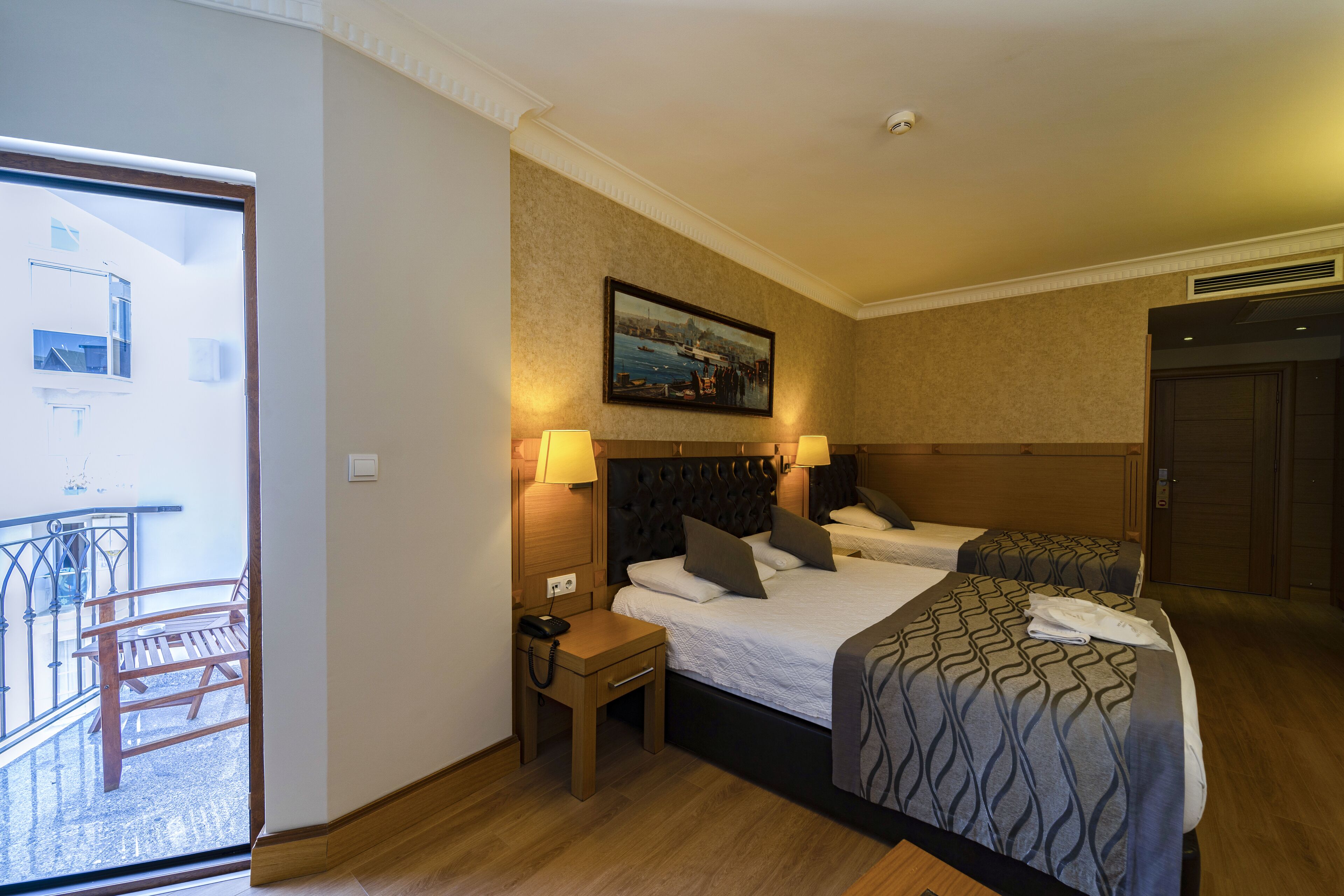 A hotel room with two beds, a small desk, a chair, and a balcony with a railing.