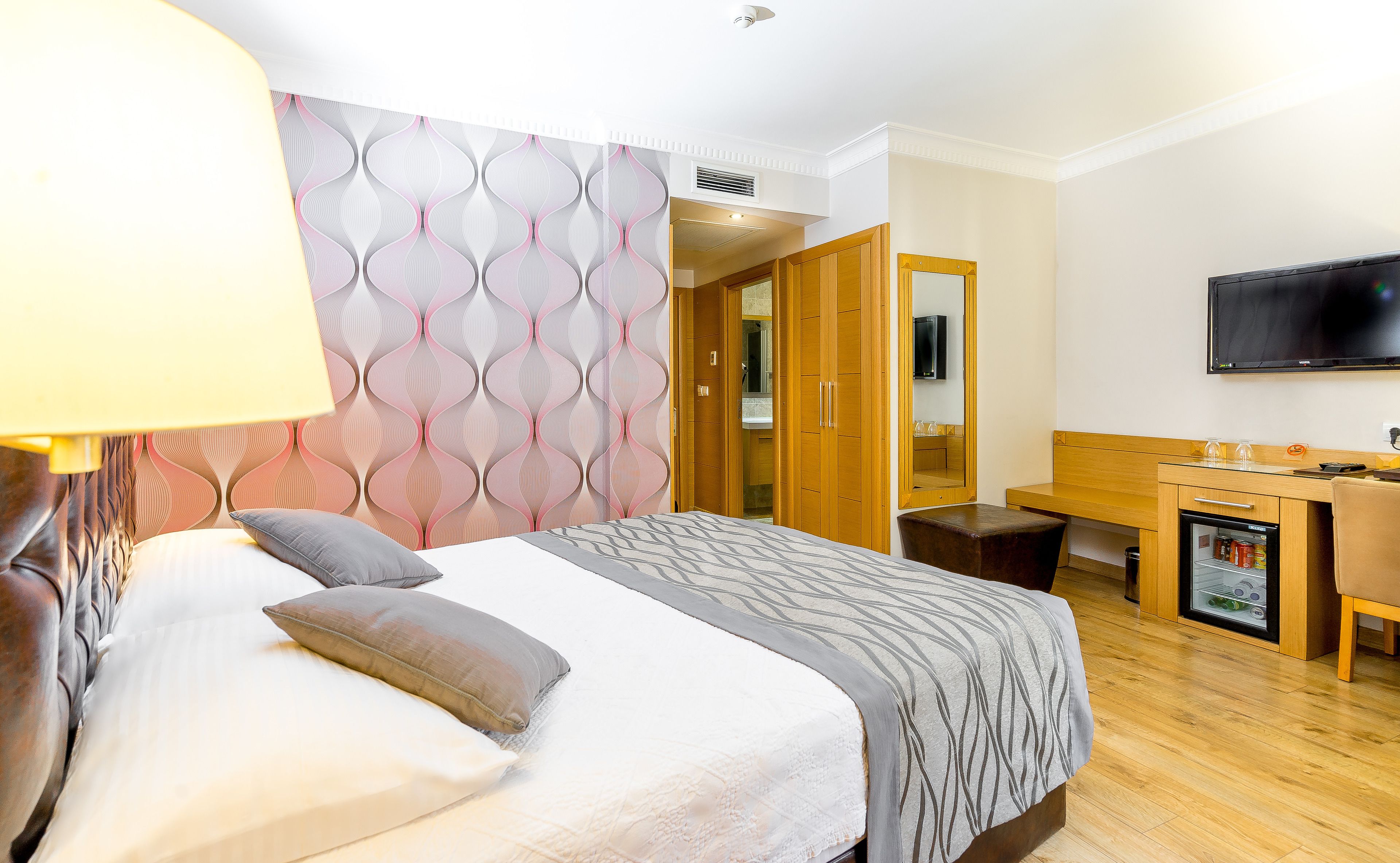 Habitación de hotel con cama, mesita de noche, lámpara, armario, escritorio, silla, televisor y un refrigerador pequeño.