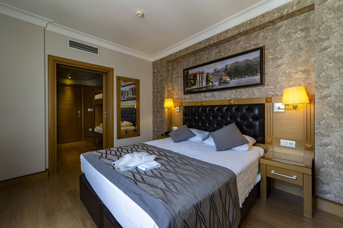 Una habitación de hotel con una cama grande, un cabecero con diseño acolchado, dos lámparas de noche, una mesita de noche de madera y un cuadro en la pared.